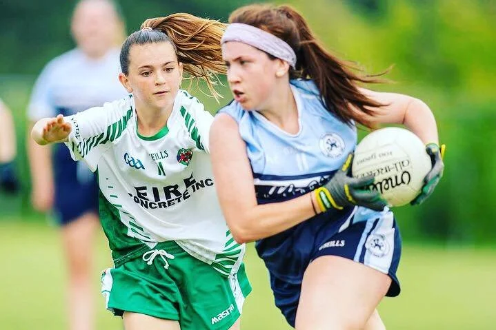 Two young girls playing Gaelic football outdoors, one in a blue uniform holding a football, the other in a green and white uniform trying to catch her.
