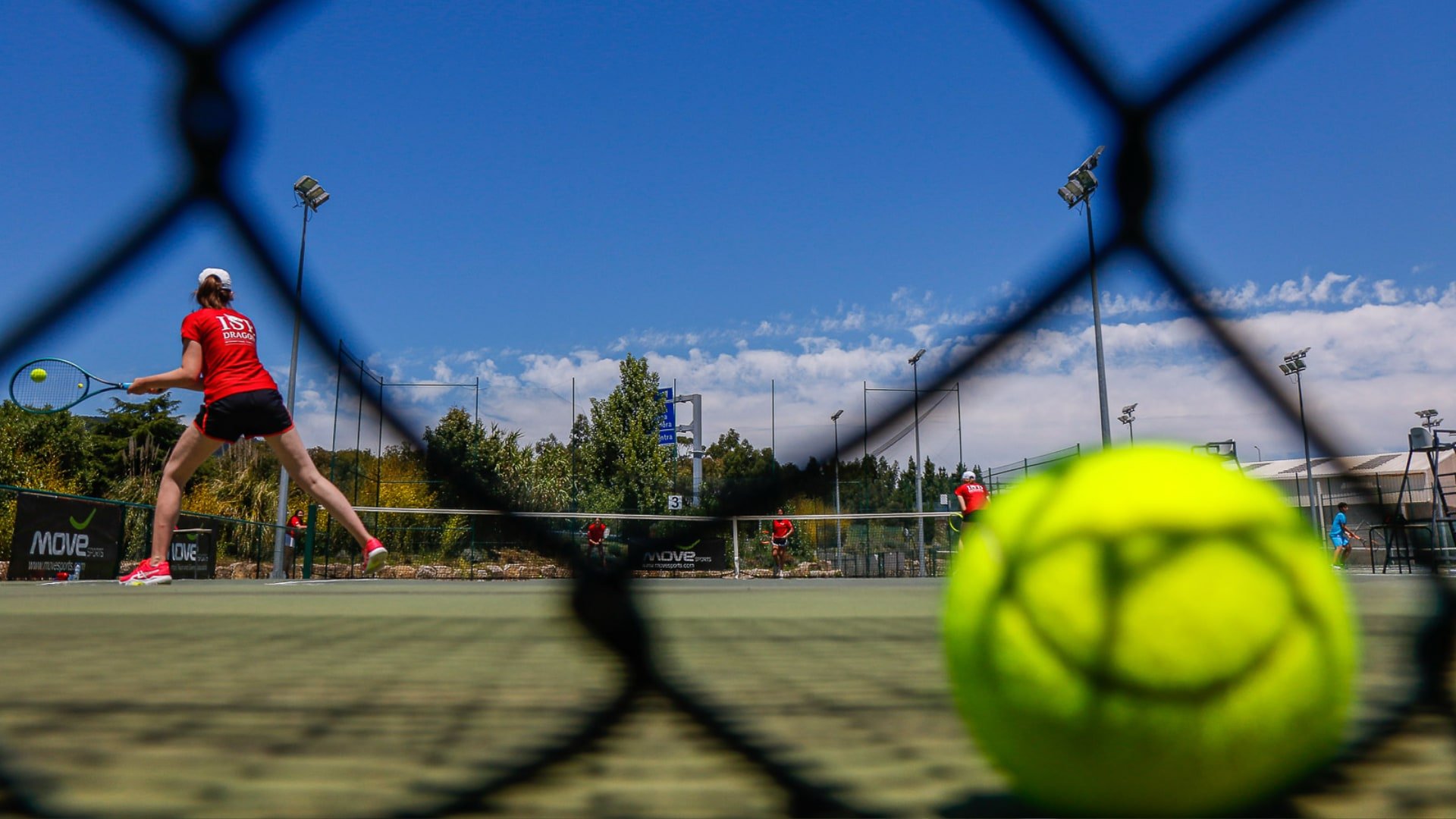 A tennis player in a red shirt and black shorts on a tennis court hitting a forehand shot. The shot is taken from behind the tennis net, with a tennis ball in the foreground and other players visible in the background under a bright blue sky.