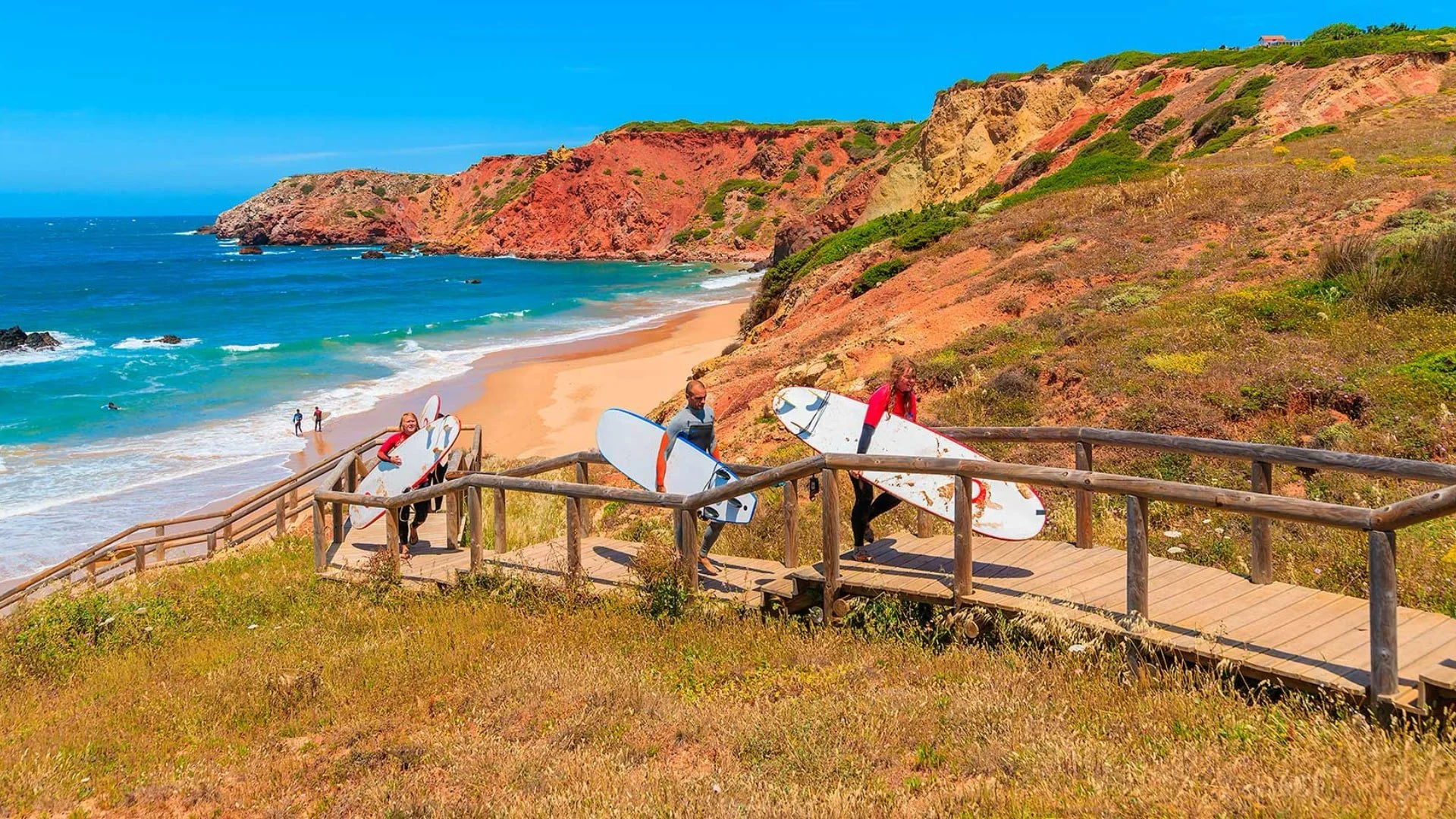 People walking with surfboards on a wooden pathway leading to a beach with cliffs and ocean in the background.