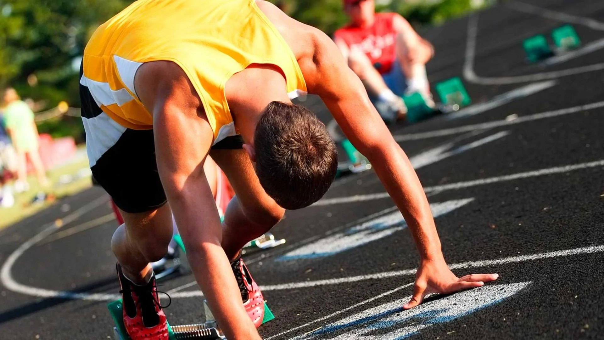 A male athlete in a yellow tank top and black shorts is in starting position on a track, preparing for a sprint race. The photo captures him from a side angle, focusing on his bent posture and hand on the ground.