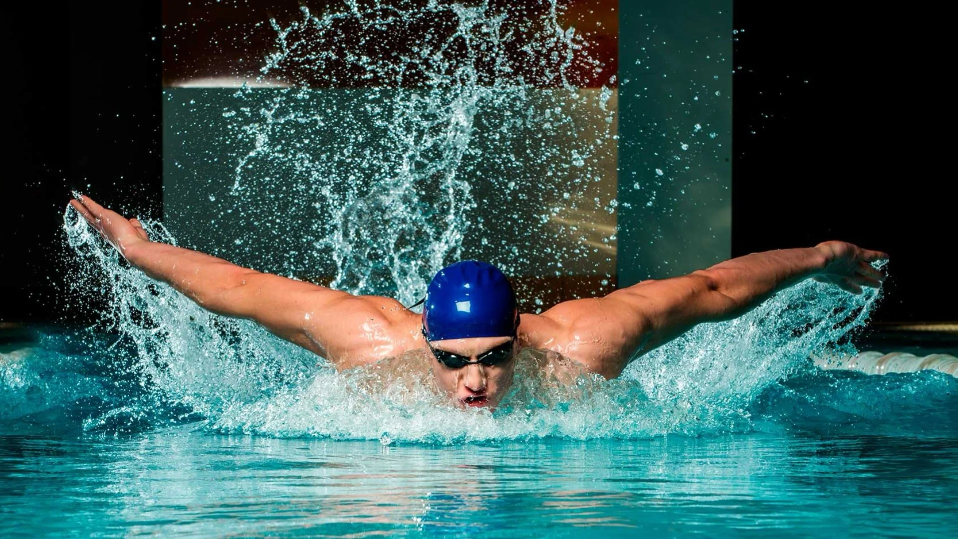 A swimmer is performing the butterfly stroke in a pool, wearing a blue swim cap and goggles, with water splashing around.
