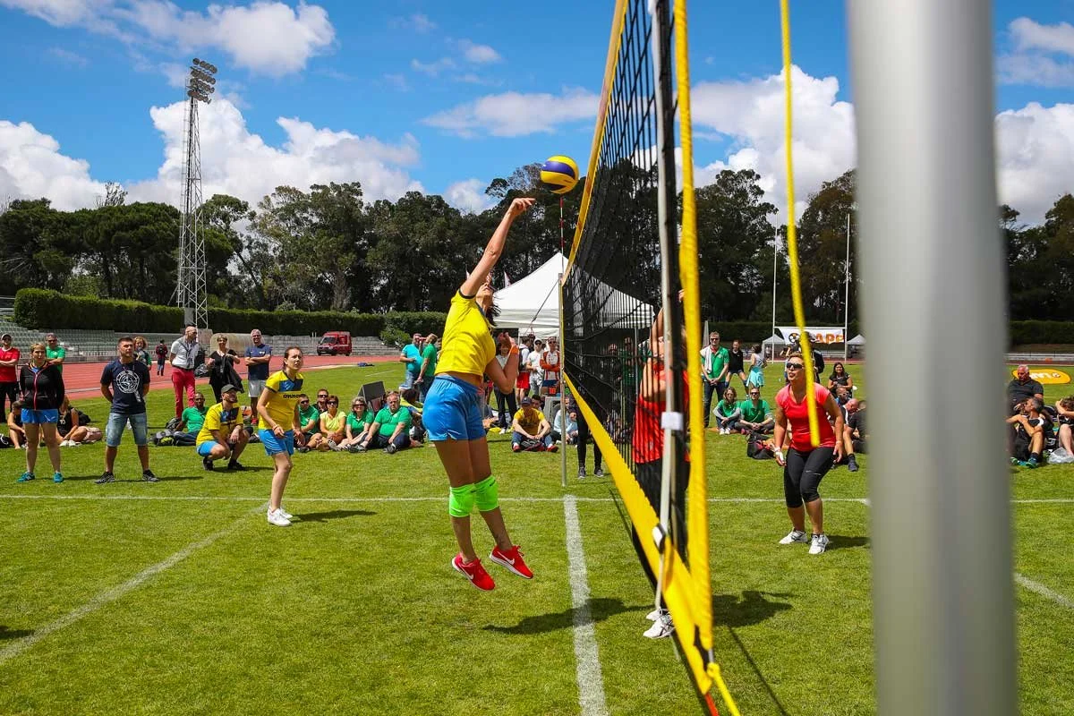 Two women playing beach volleyball on a grassy field, with one jumping to hit the ball over the net. There are spectators sitting and standing around the court, and other players and officials in the background. Clear blue sky with a few clouds.