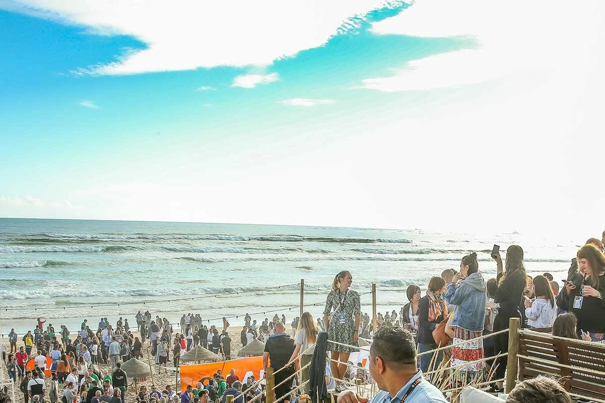 Crowded beach scene with many people, some taking photos, overlooking the ocean on a sunny day with blue sky and clouds.