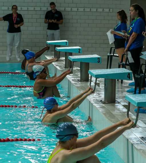 Swimmers at a pool starting block during a race, with officials and coaches watching.