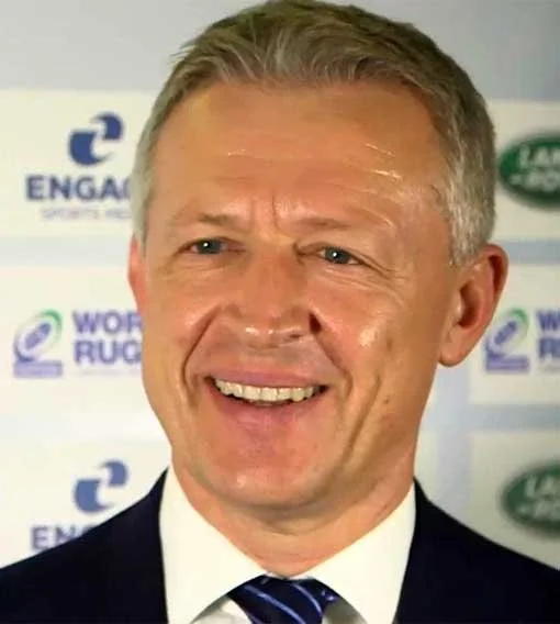 A smiling blonde man wearing a dark suit, white shirt, and striped tie, standing in front of a backdrop with logos of Land Rover, World Rugby, and Engaget