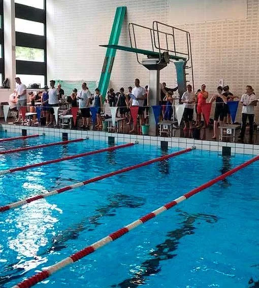 Swimmers waiting in line at an indoor swimming pool facility with blue and white lane dividers, poolside seating, and a high diving platform.