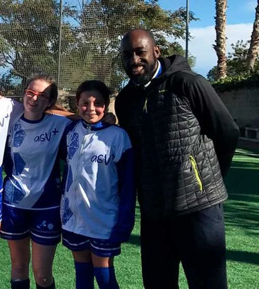 Two young female soccer players in blue and white uniforms standing with a male coach on a soccer field outside, with trees and a clear sky in the background.