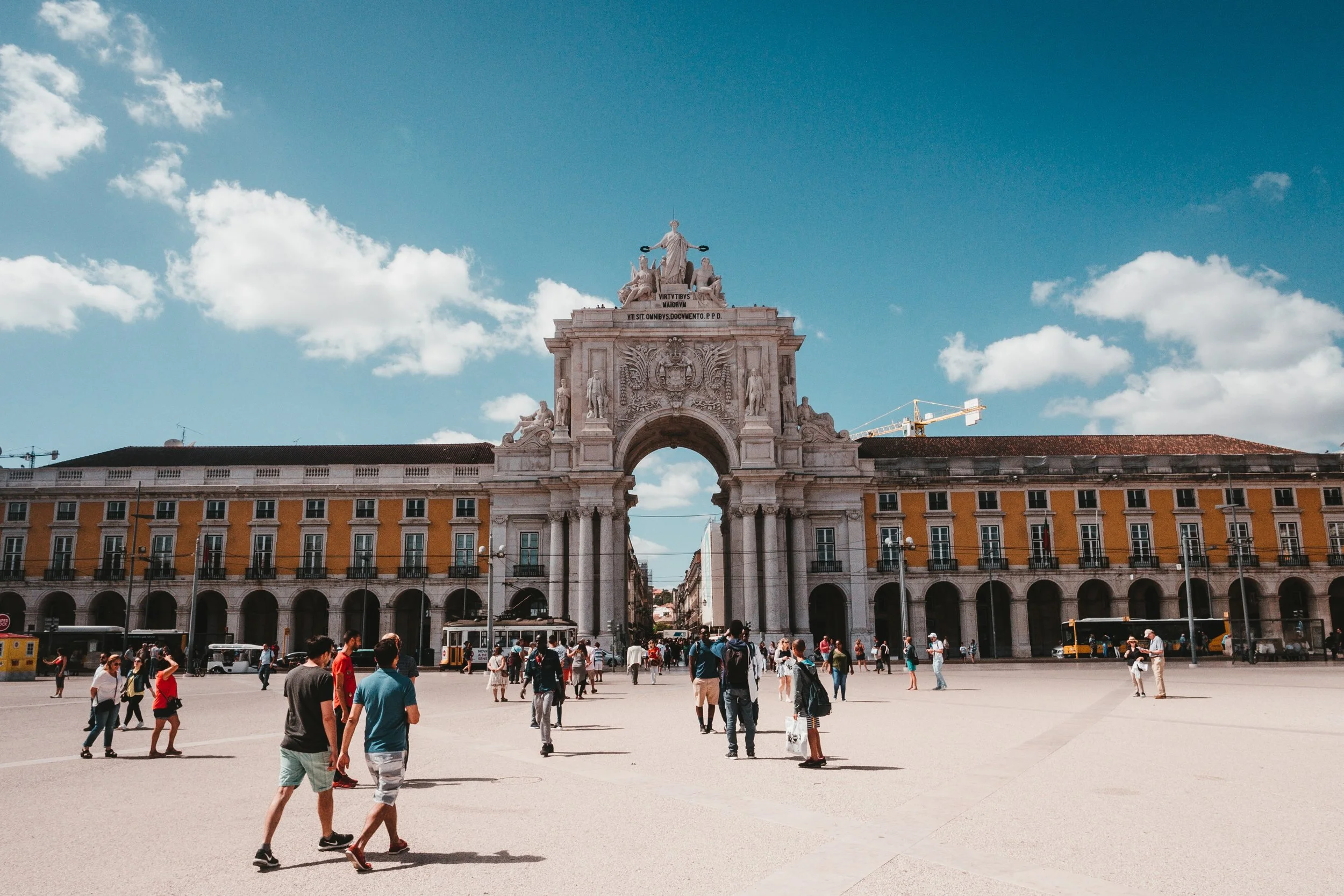 People walking in a large open plaza with a grand archway and historical buildings in the background, under a partly cloudy sky.
