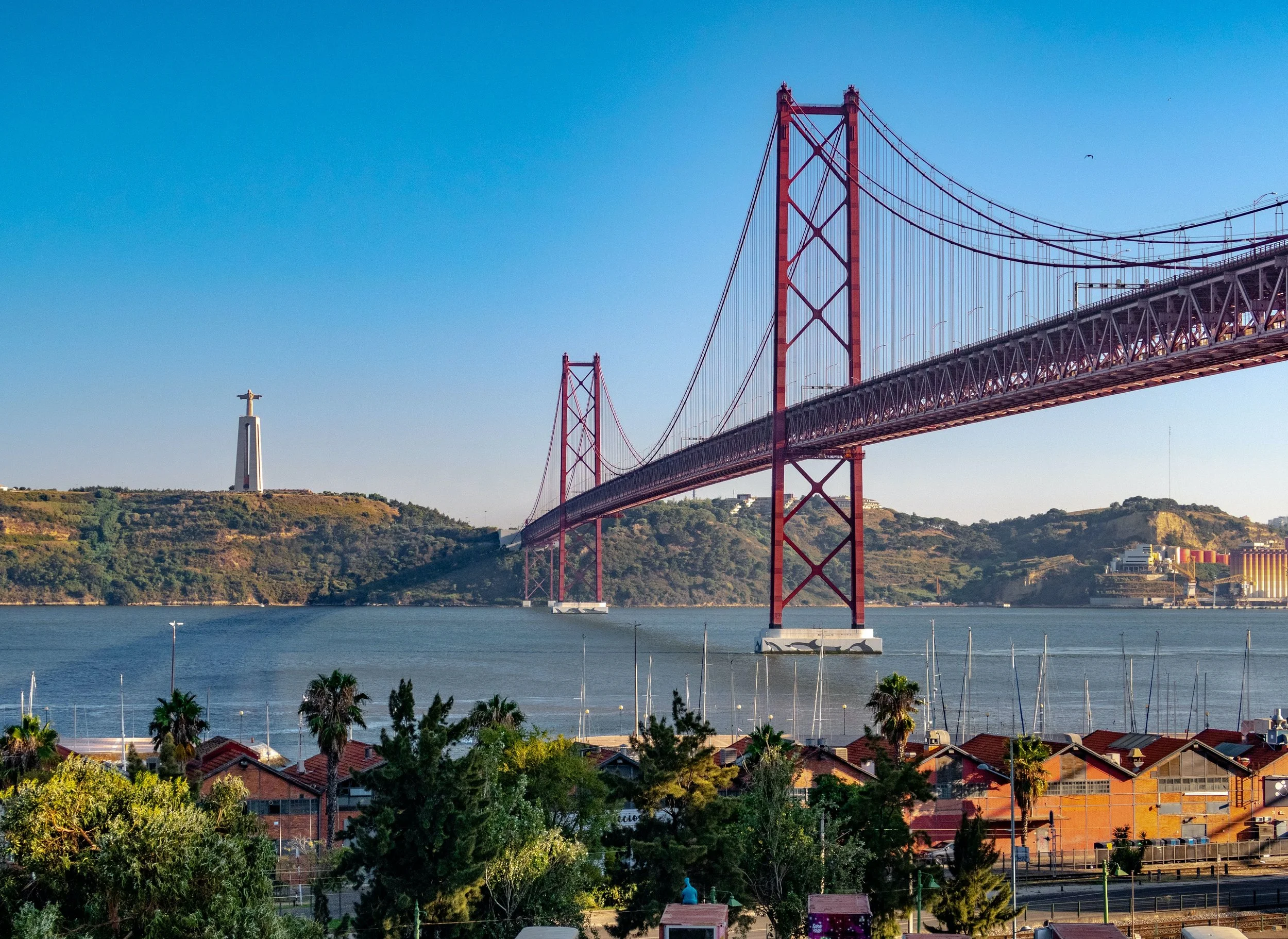 View of the Golden Gate Bridge in San Francisco, California, with the Christ the King statue on a hill in the background, and a marina with sailboats in the foreground.