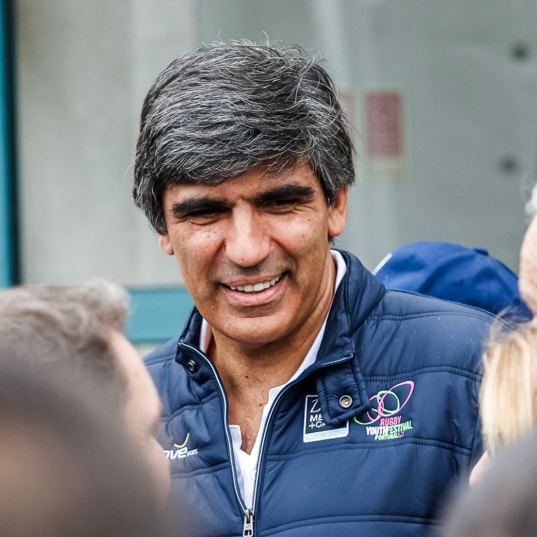 A man with dark gray hair smiling and talking to children outdoors, wearing a blue sports jacket with event logos.