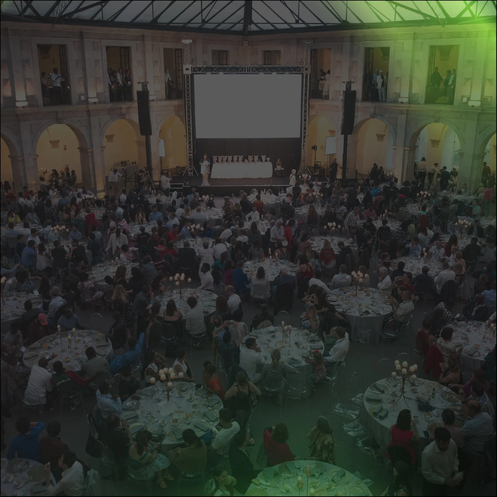 A large banquet hall filled with round tables set for a wedding reception or event, with many guests seated and standing. There is a stage with people standing and a large screen displaying a video or picture of some men. The hall has high ceilings, columns, and balconies overlooking the main floor.