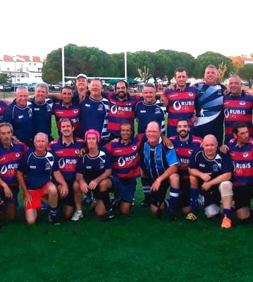 Group of rugby players posing on a field, some standing and some kneeling, with goalposts and trees in the background, wearing team jerseys with sponsor logos.