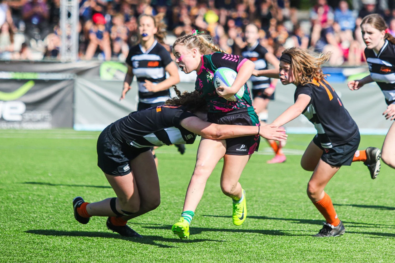 Girls’ rugby match action during the Portugal Rugby Youth Festival, with a player breaking a tackle while carrying the ball on a grass pitch.