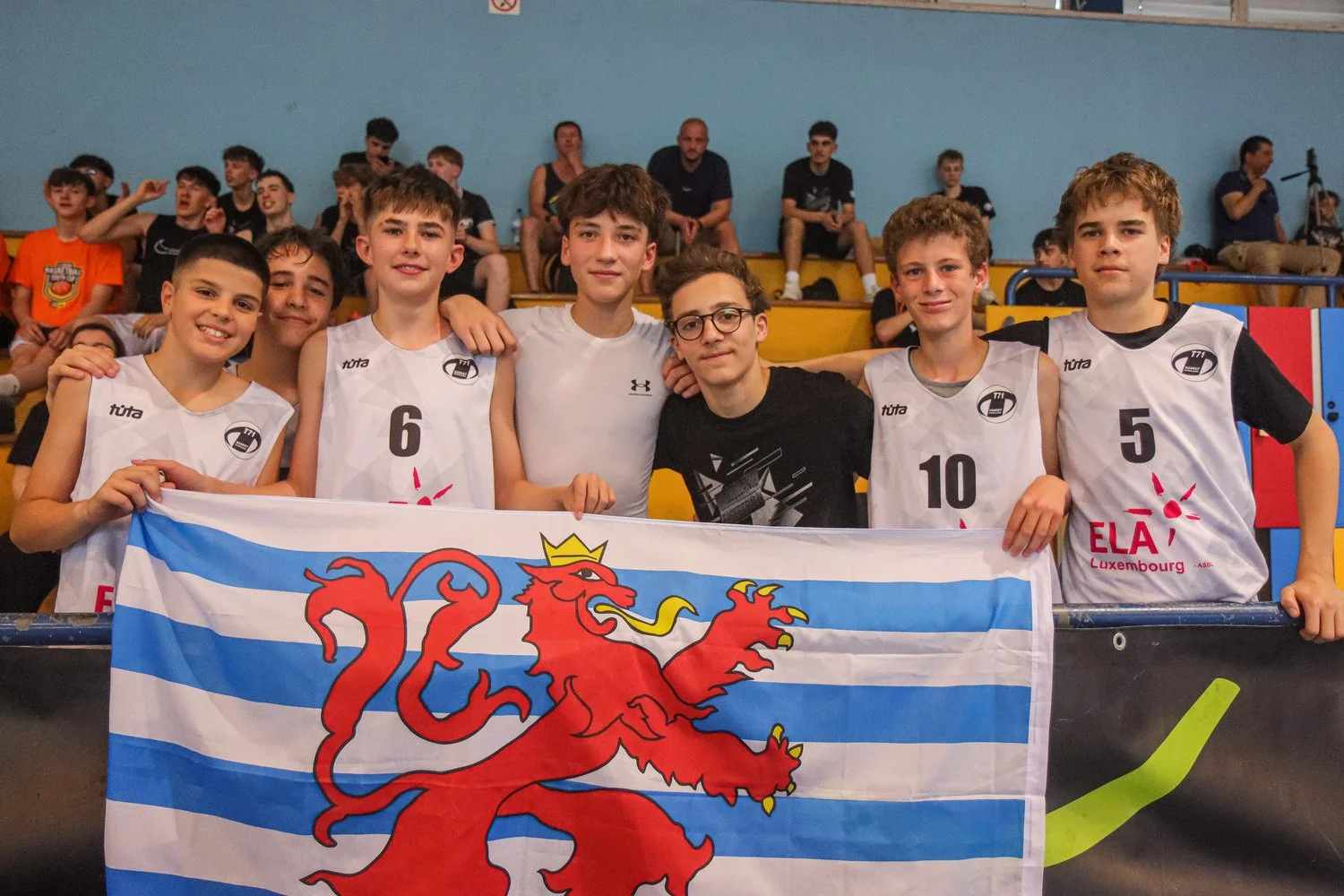 Youth basketball players posing with the Luxembourg flag in the stands during the Lisbon Basketball Youth Cup.