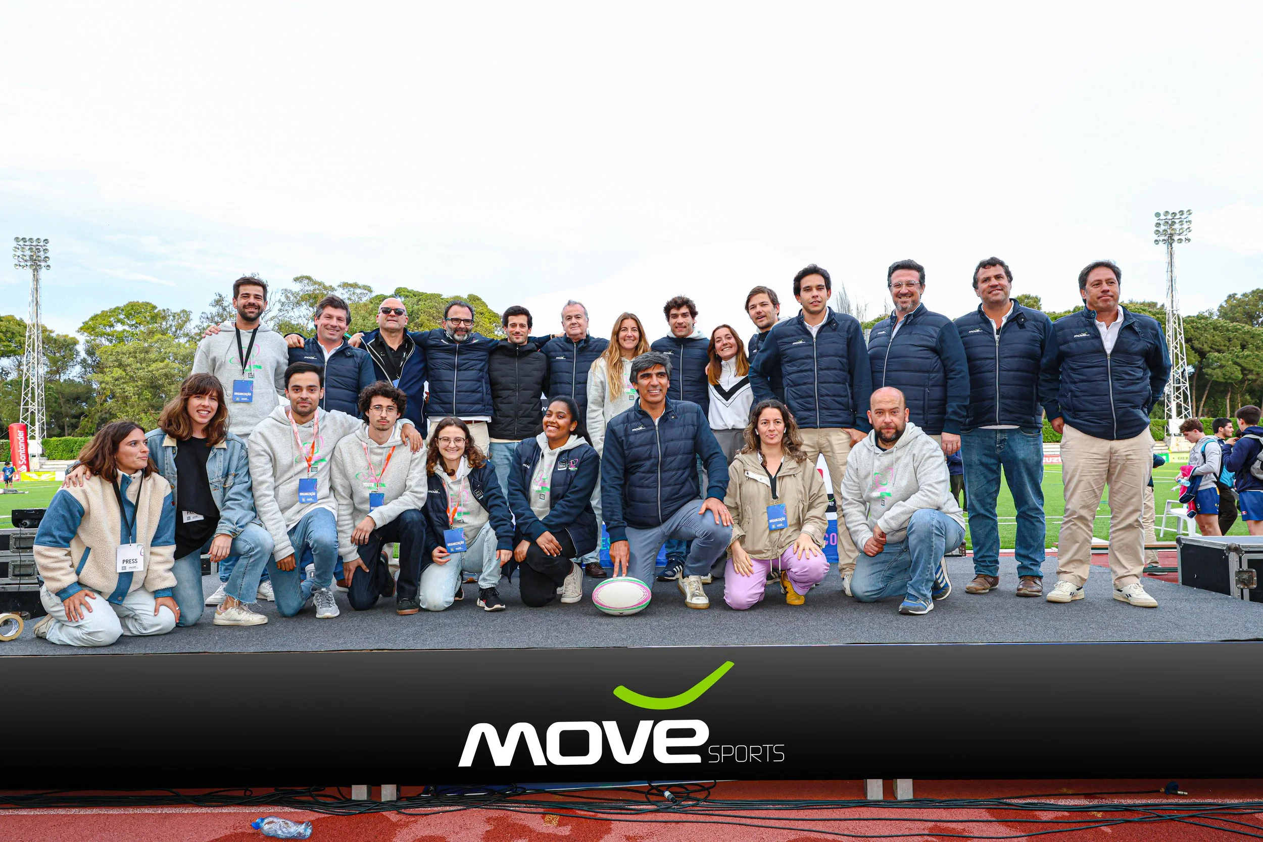 Group of people at an outdoor sports event, standing and kneeling on a stage with a 'MOVE SPORTS' banner, some wearing jackets and lanyards, with a sports field and trees in the background.