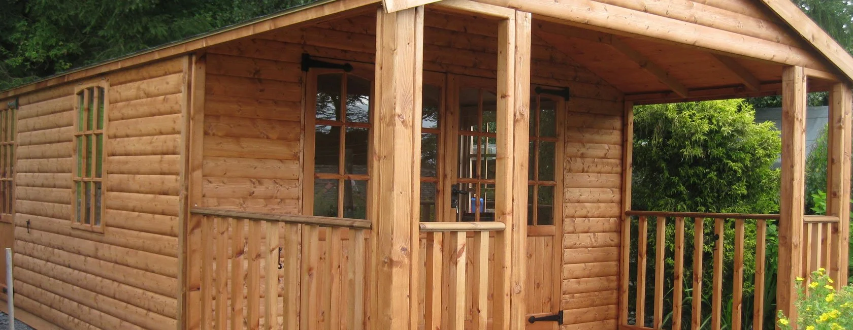 Wooden cabin with a porch and windows, located in a garden with greenery and flowers.