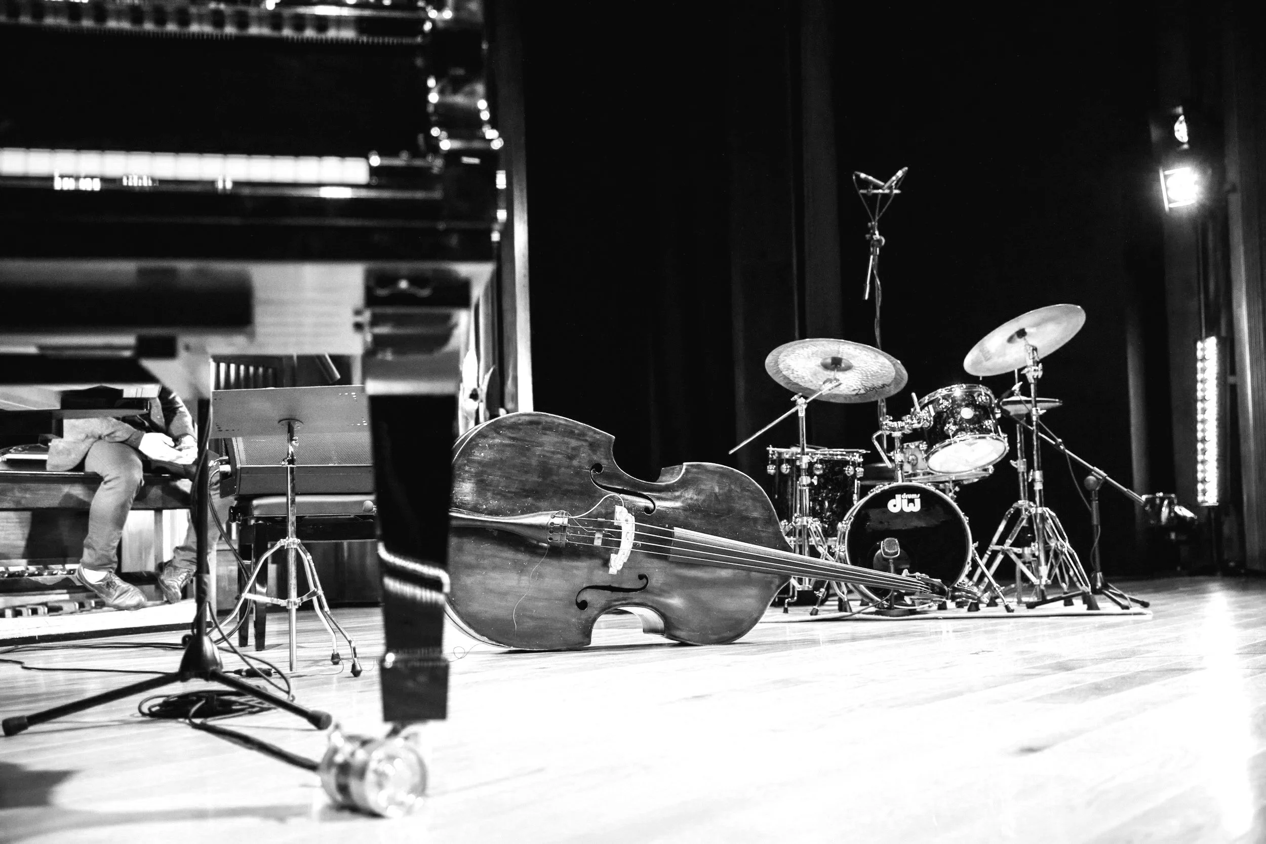 Stage with a double bass, drum set, and a person sitting at a piano in a music venue.
