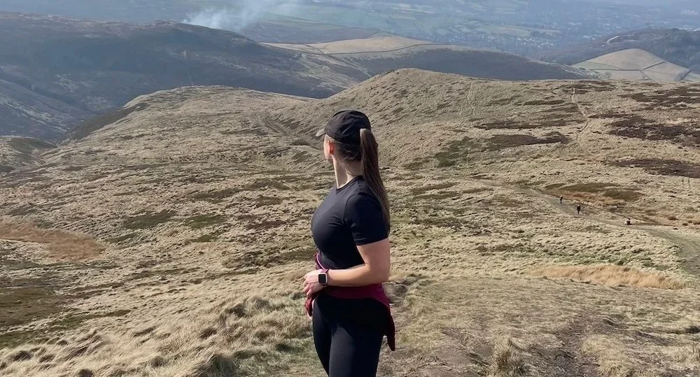 Confident life coach standing on a mountain trail in black athletic clothing and a cap, overlooking rolling hills and valleys, feeling strong, inspired, and ready for personal growth.