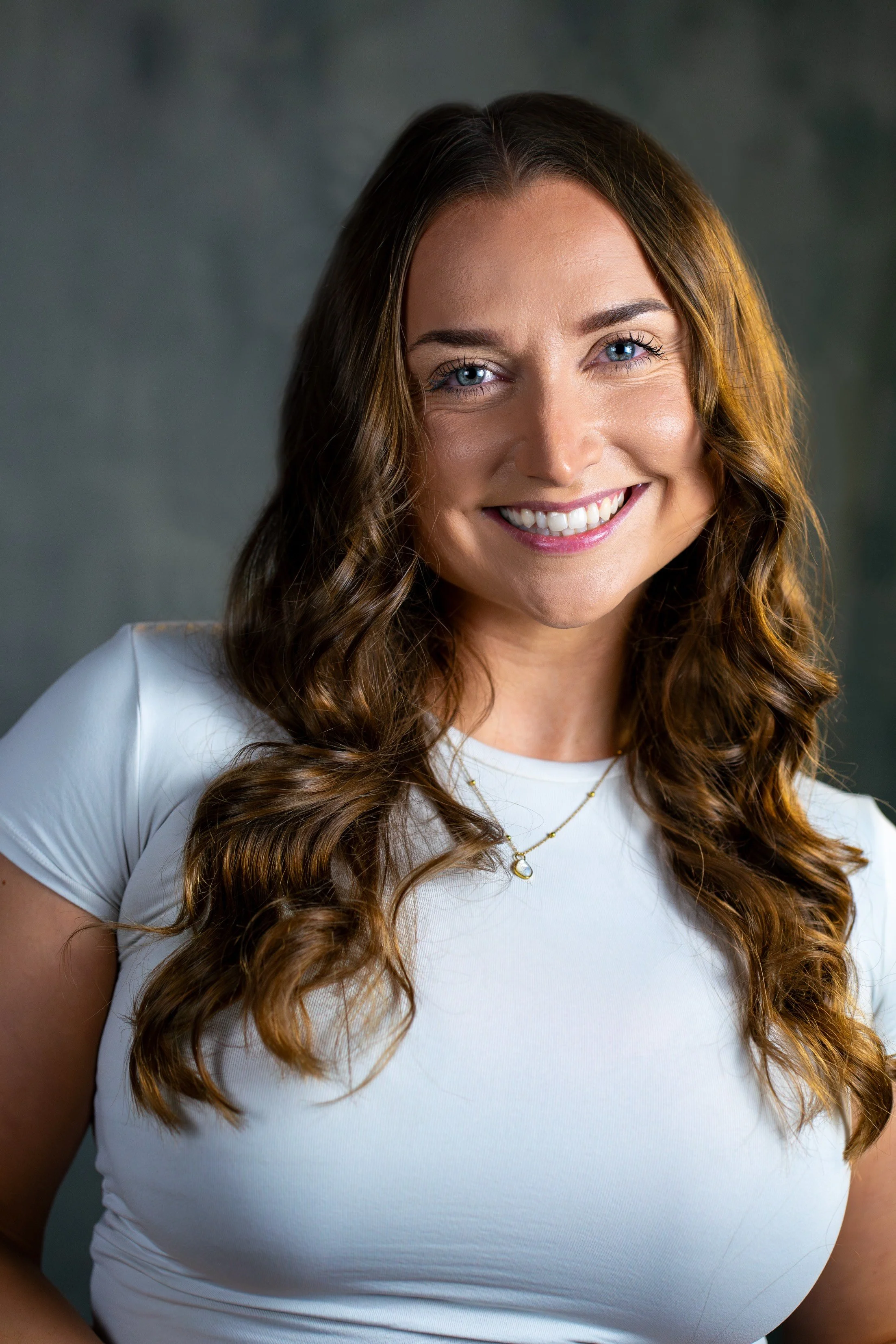 Friendly life coach with long wavy brown hair and blue eyes, smiling warmly, wearing a white T-shirt and gold necklace against a dark grey background.