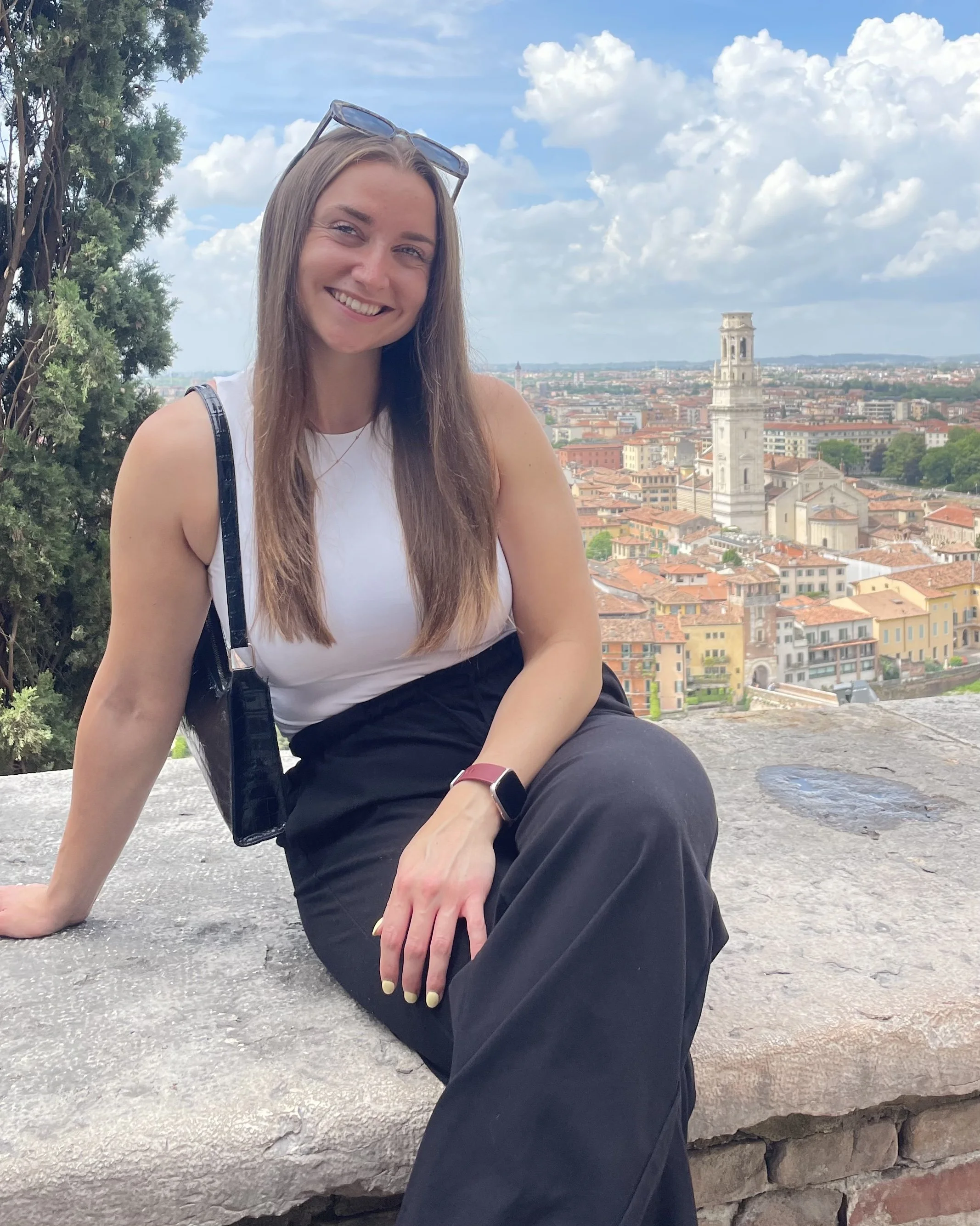 Friendly, personable life coach with long brown hair, wearing a white sleeveless top and black pants, sitting on a stone surface with a cityscape behind her under a partly cloudy sky.