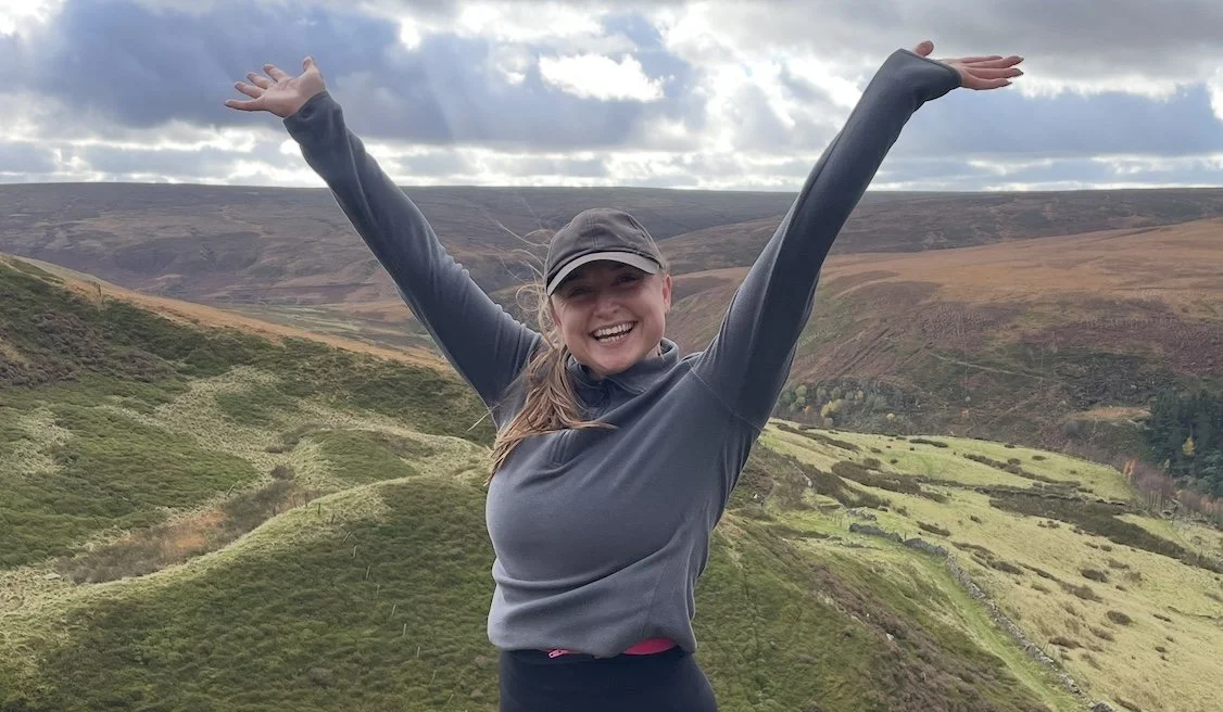 Joyful life coach smiling and raising her arms in a scenic outdoor landscape with rolling hills and a cloudy sky, feeling empowered, free, and inspired.