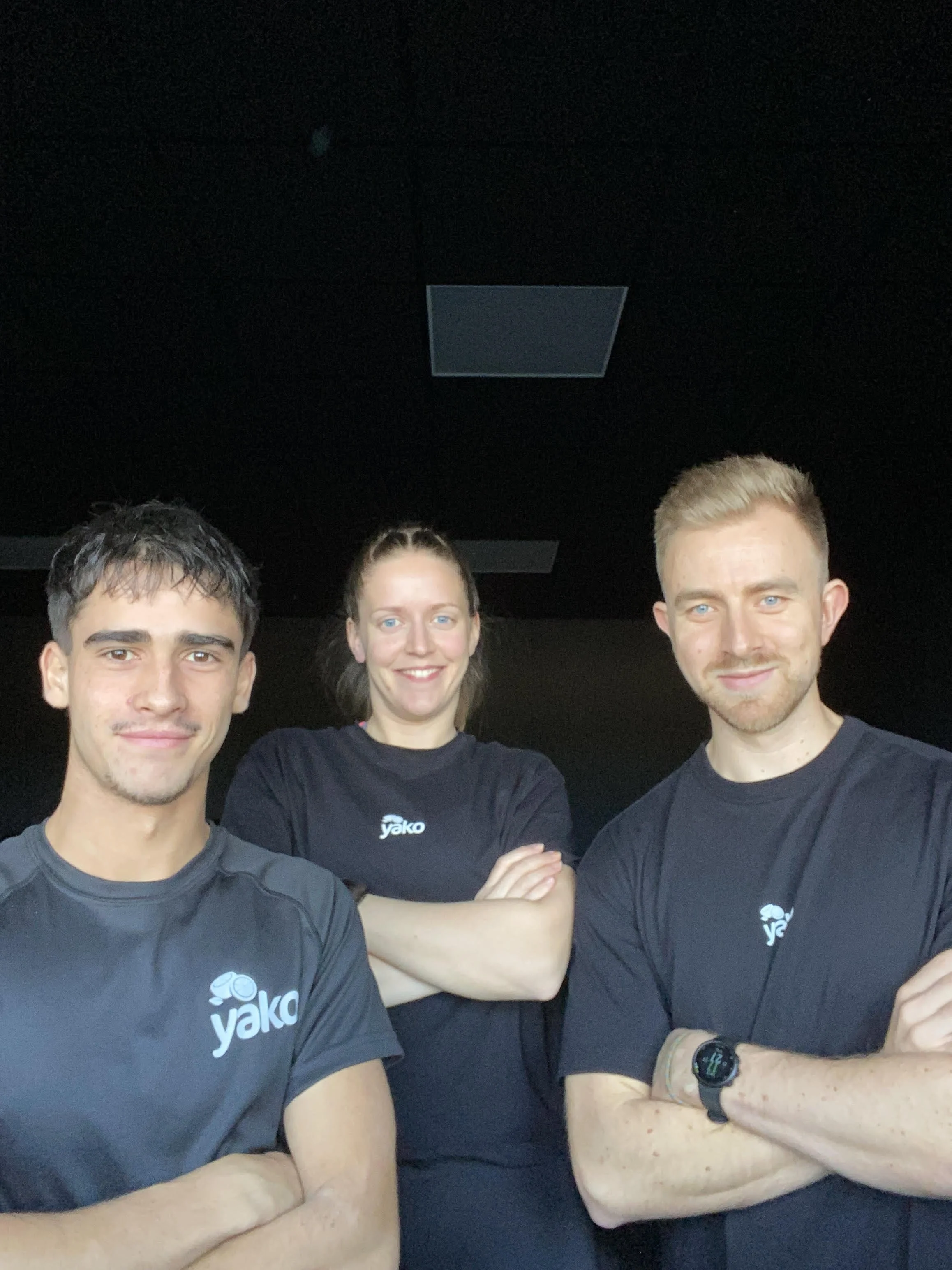 Three young adults standing against an orange background, wearing casual and athletic clothing, with arms crossed and smiling.