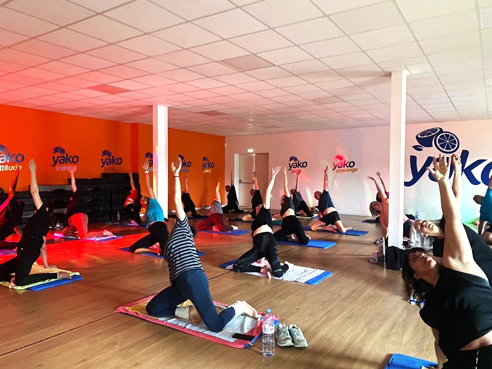 Group of people participating in a yoga or stretching class in a brightly lit studio, some on their knees with one arm extended upward, on blue yoga mats, with orange and white walls displaying the Yako logo and words like training, jump, pump, challenge.