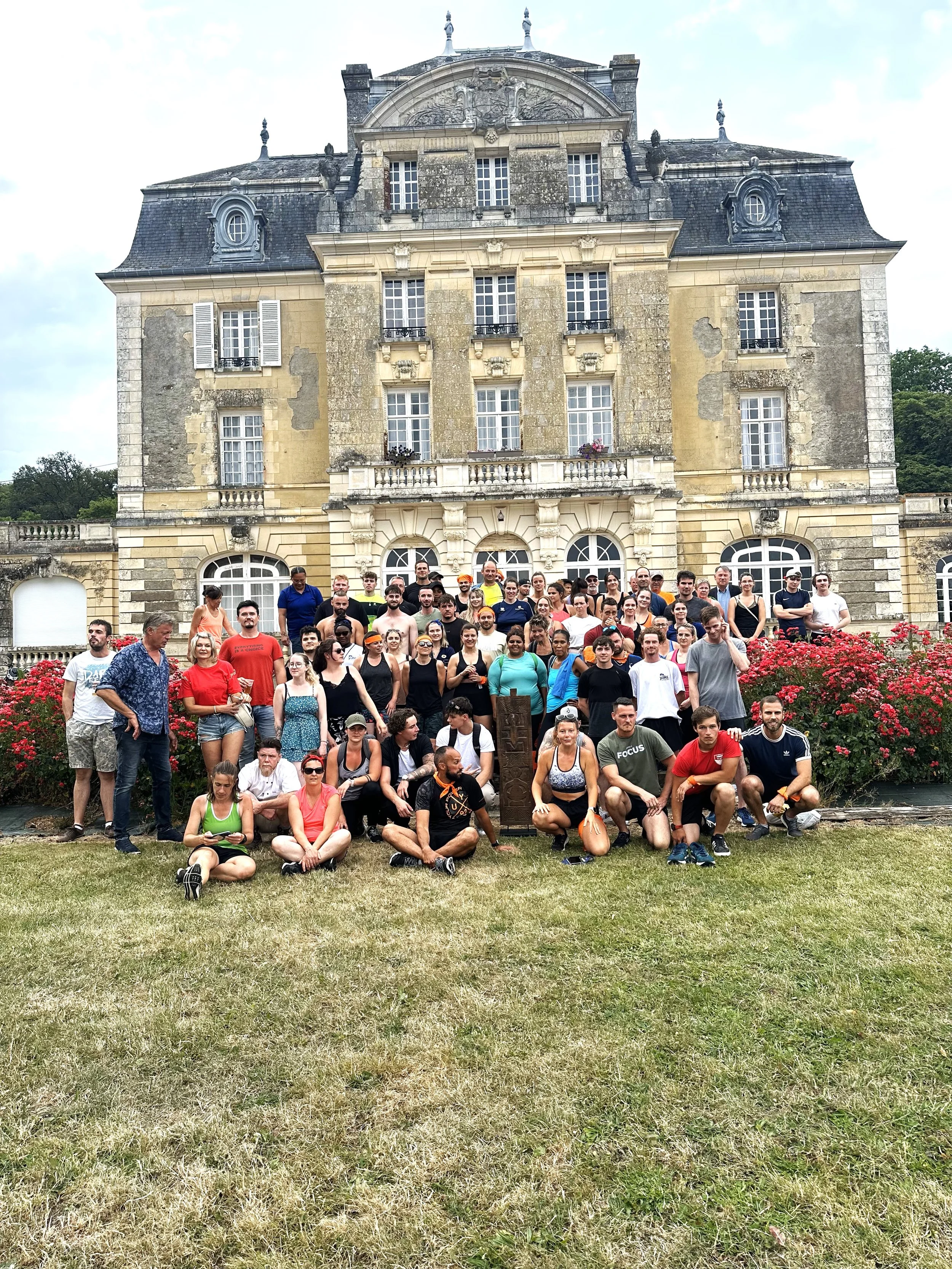 A large group of people posing in front of a historic stone building with a landscaped garden and blooming flowers.