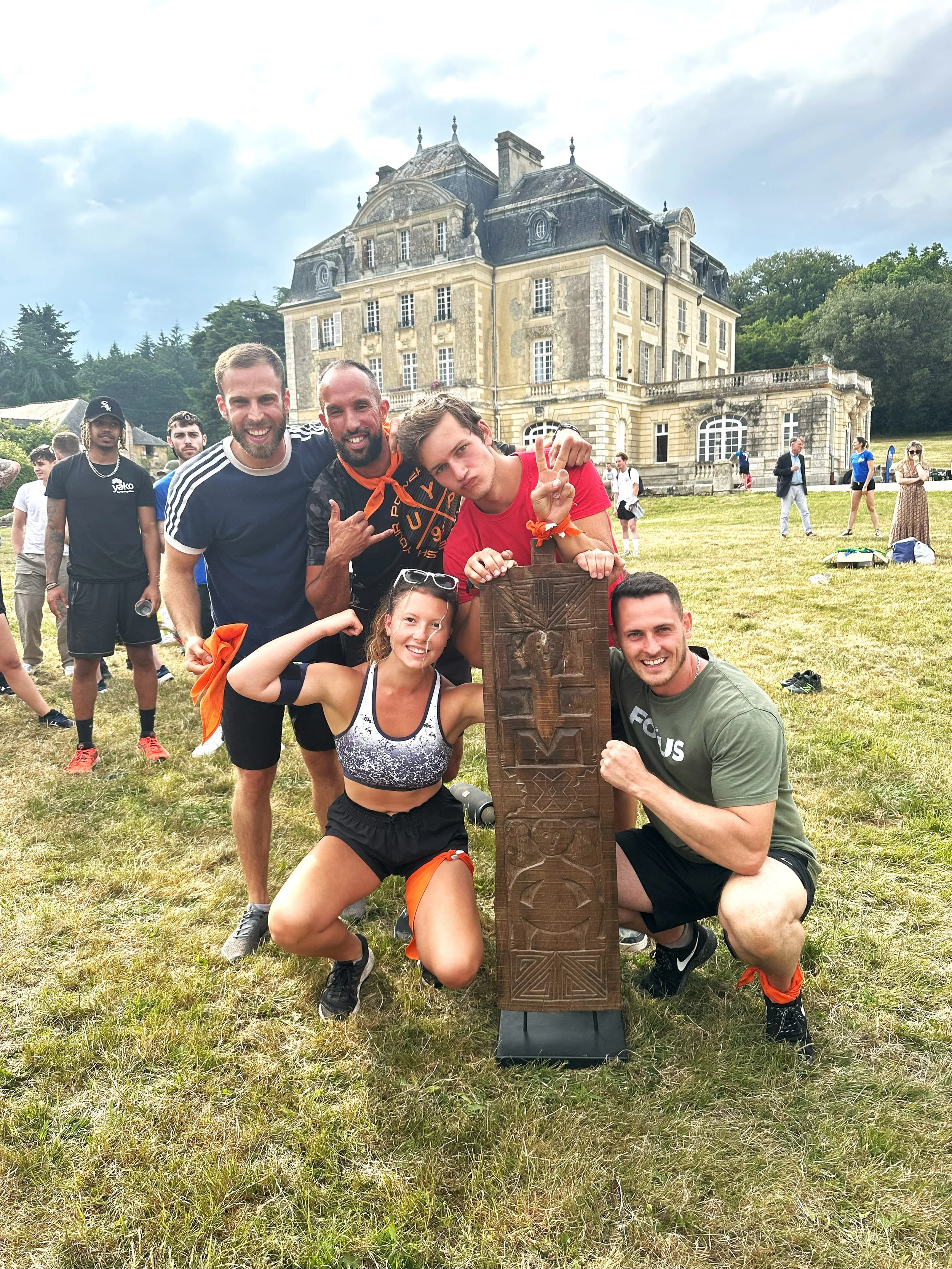 Group of six excited people outdoors, posing with a carved wooden artifact in front of a historic castle-like building on a grassy field.