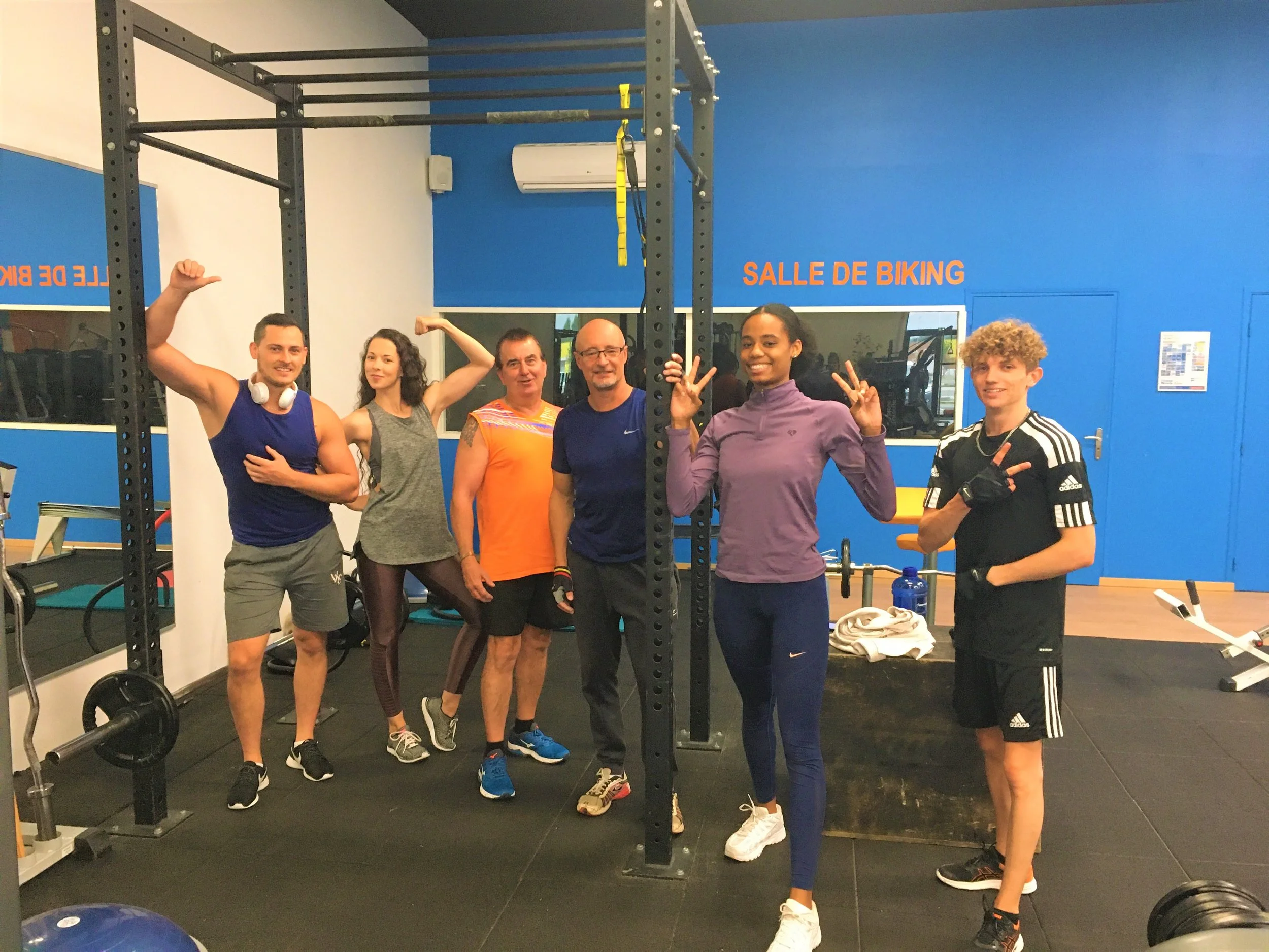 Group of seven people posing inside a gym with weightlifting equipment, some making poses, and a sign that reads 'SALLE DE BIKING' on the wall.