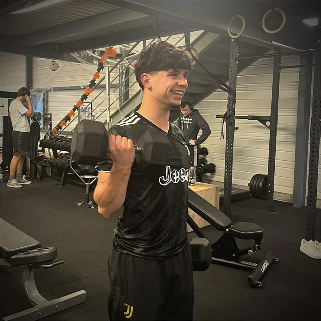 Man smiling and exercising on a fitness machine at a gym, with a woman working out in the background.