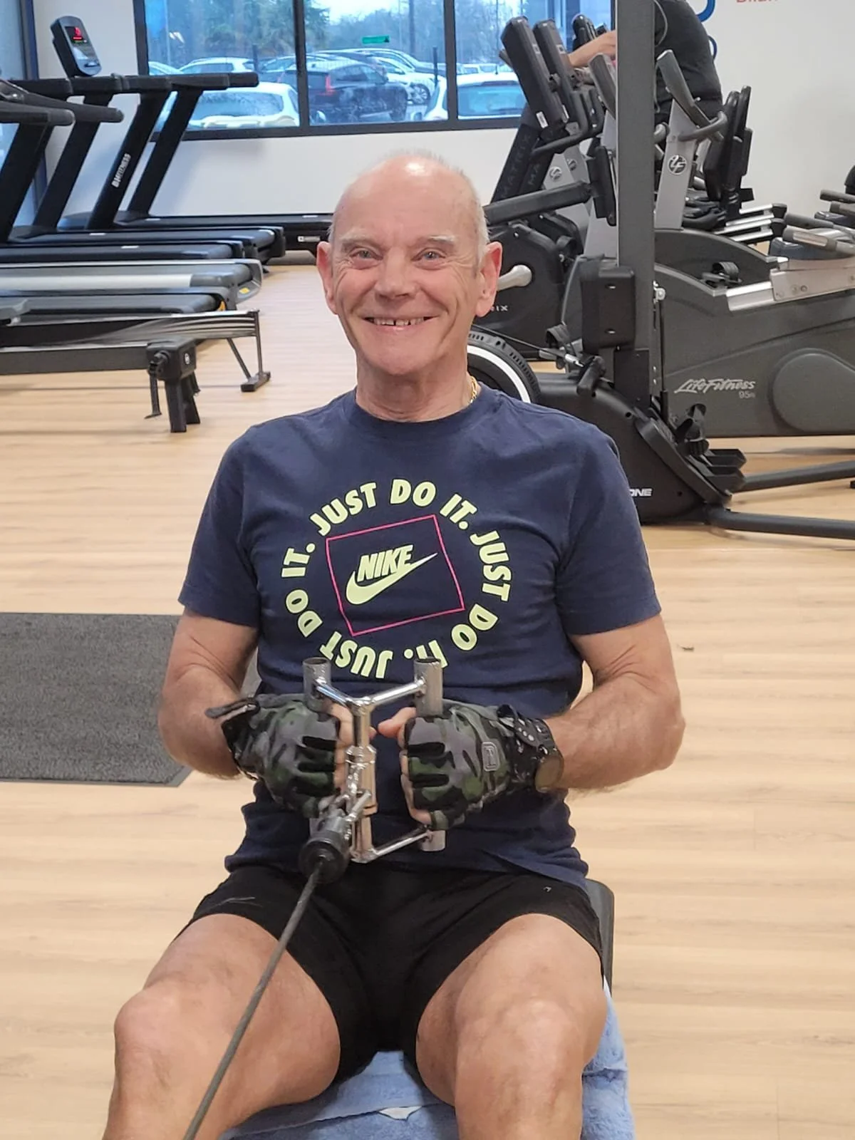 Man smiling and exercising on a fitness machine at a gym, with a woman working out in the background.