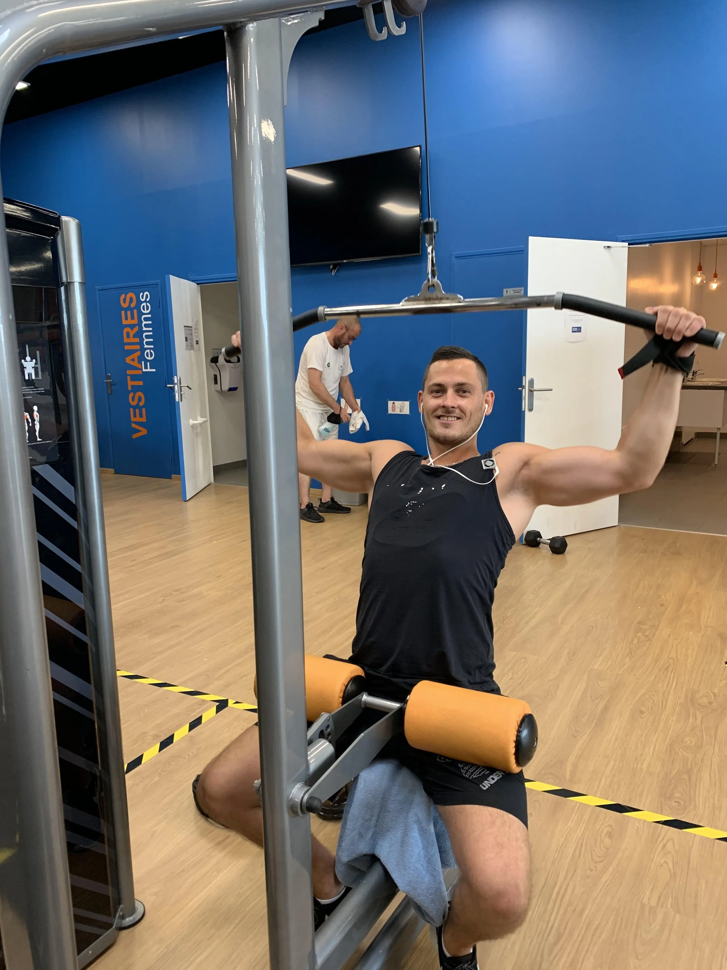Man smiling and exercising on a fitness machine at a gym, with a woman working out in the background.