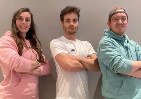 Three young adults standing against an orange background, wearing casual and athletic clothing, with arms crossed and smiling.