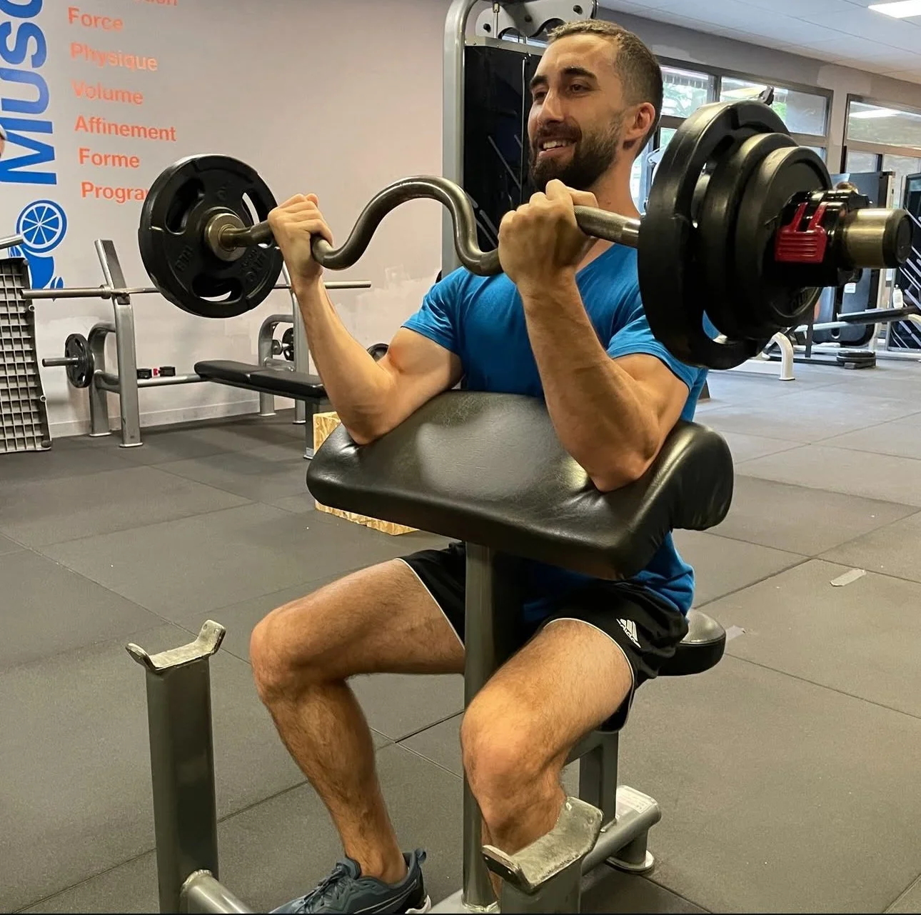 Man smiling and exercising on a fitness machine at a gym, with a woman working out in the background.