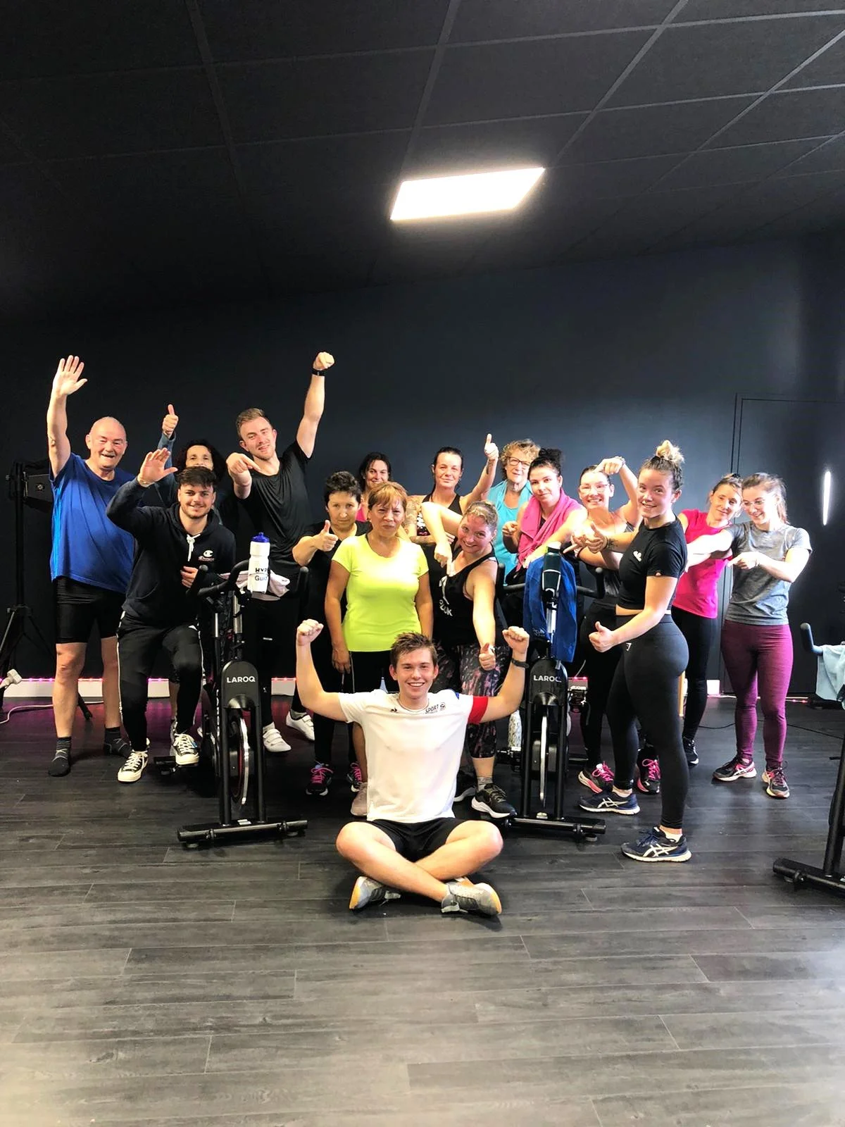 Group of diverse people, including men and women, gathered in a gym, celebrating after a workout with smiles, some giving thumbs up, and some flexing their muscles.
