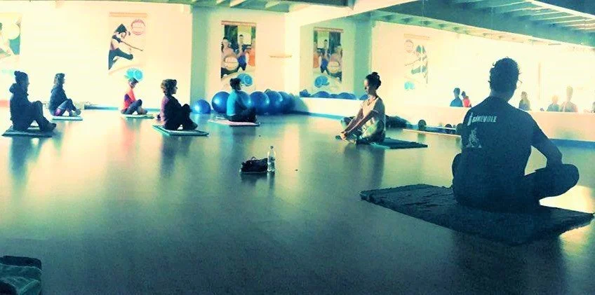 People participating in a yoga class in a bright, spacious studio with large windows and exercise balls along the wall.
