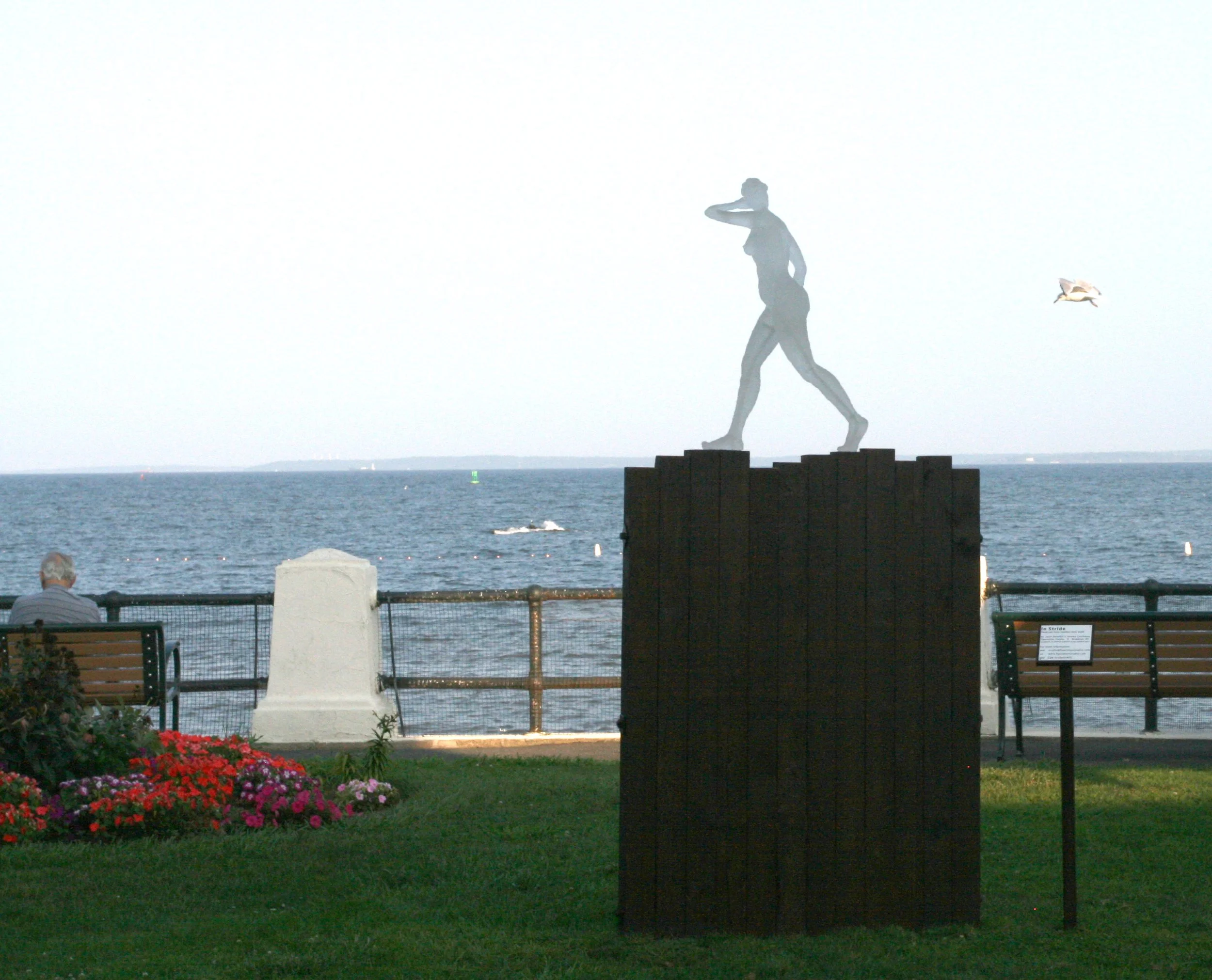 Statue of a woman walking on a platform near the water with a bird flying nearby, benches, flowers, and a fence in the foreground.