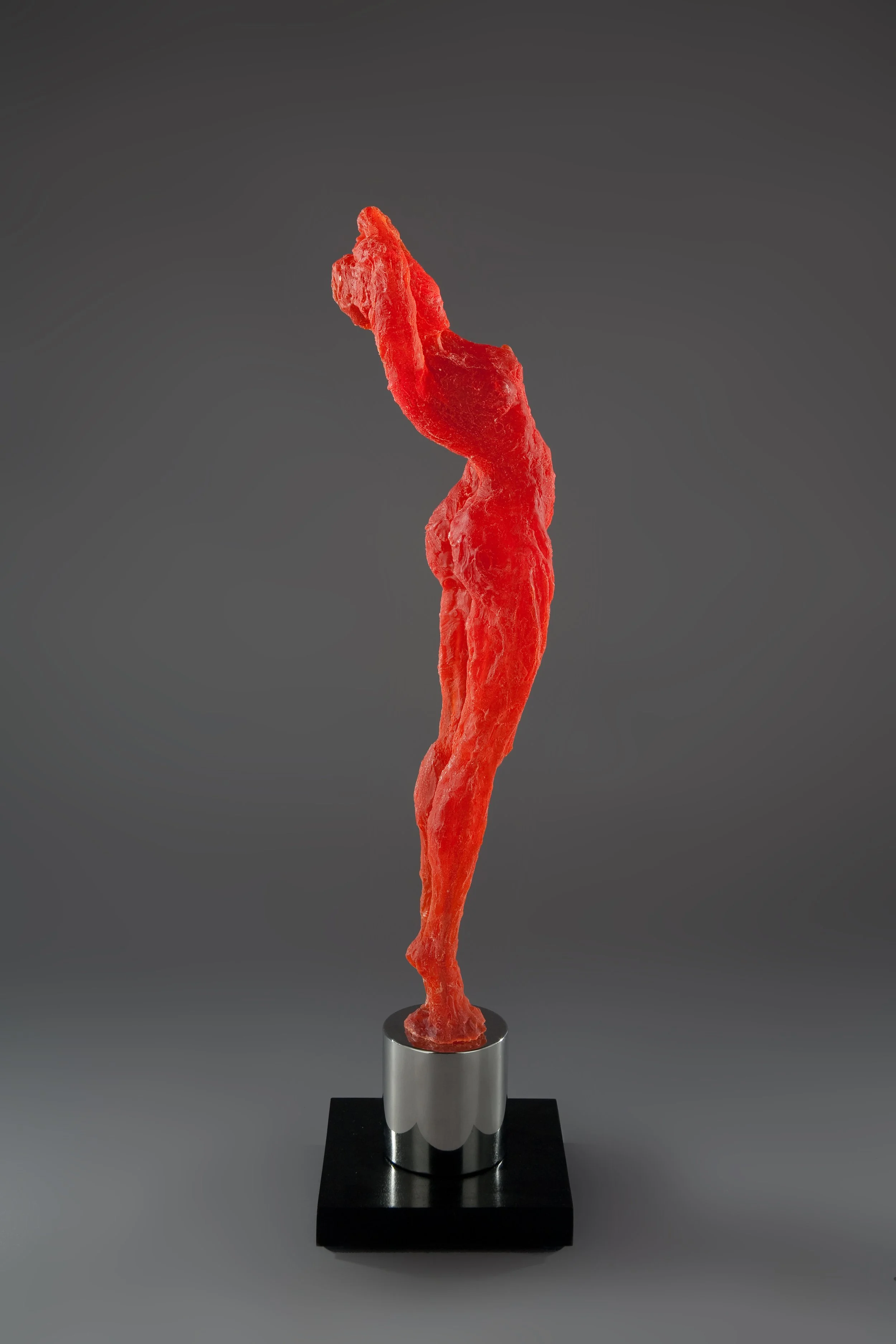 A glowing, luminous sculpture of a orange-red, elongated, and abstract female figure standing arching upwards on a black base with a polished stainless steel column.