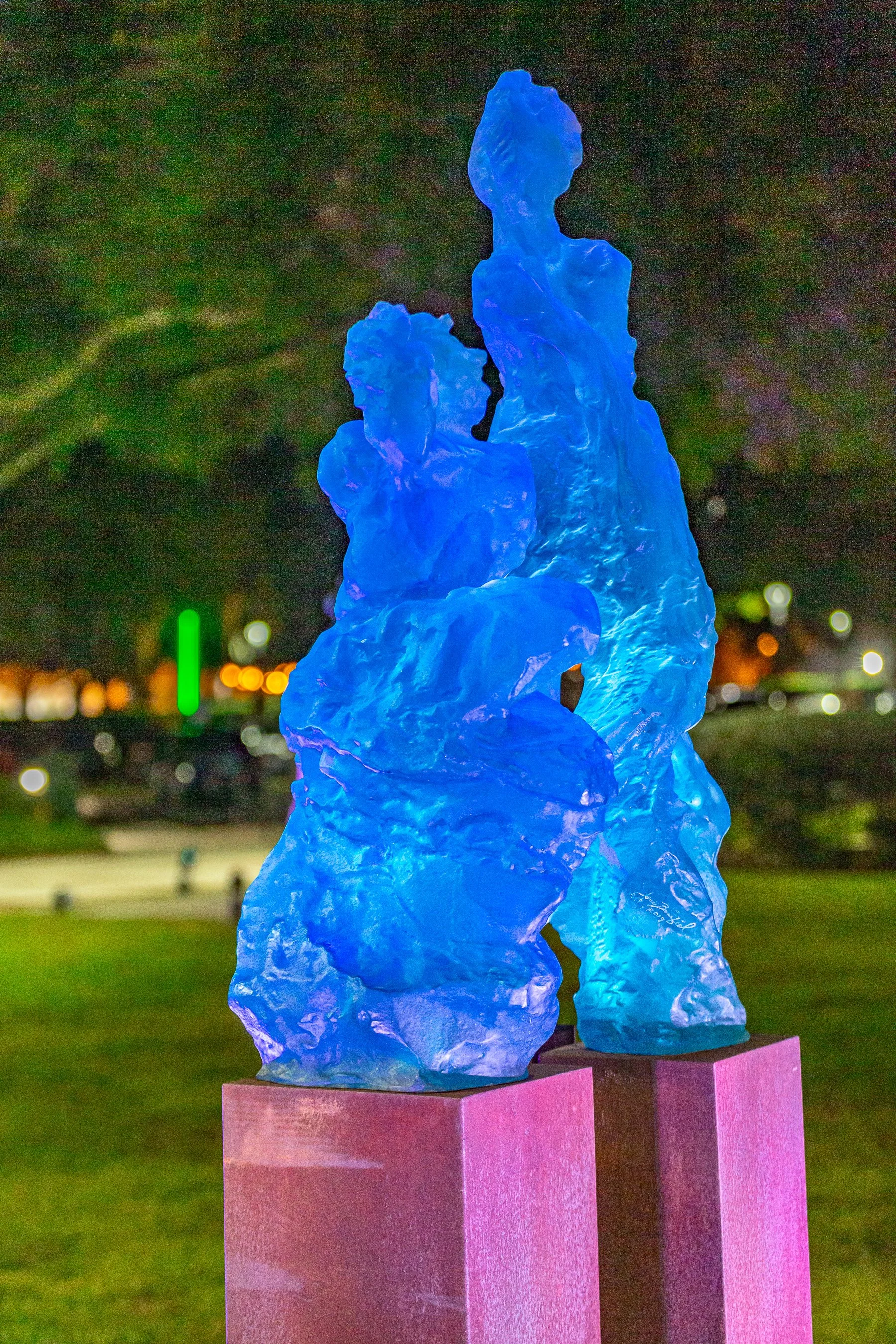 Blue abstract sculpture illuminated at night, placed on purple pedestal in a park with faint city lights in the background.