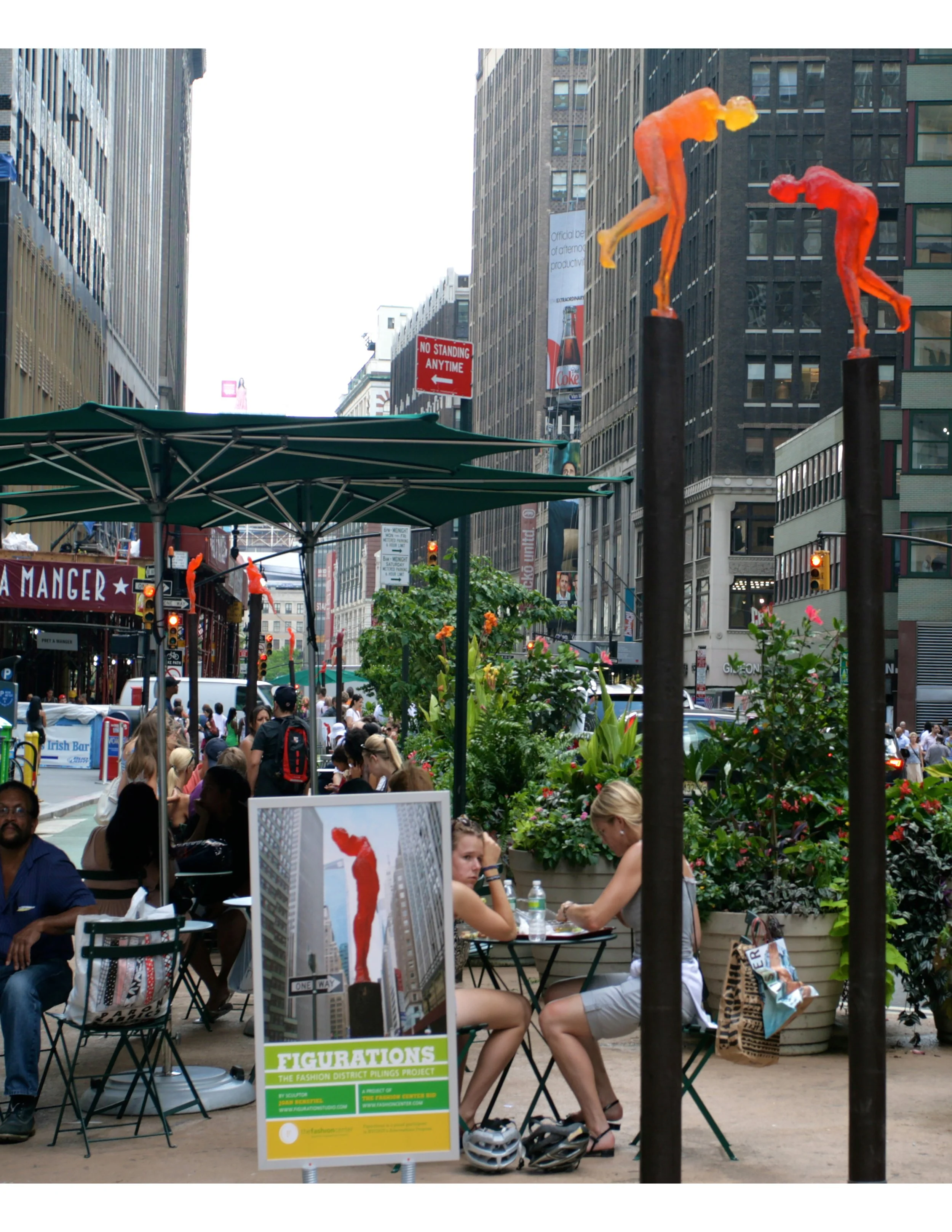 People sitting at tables and walking in a city street, with large sculptures of human figures on black poles, and a sign advertising a fashion district project.