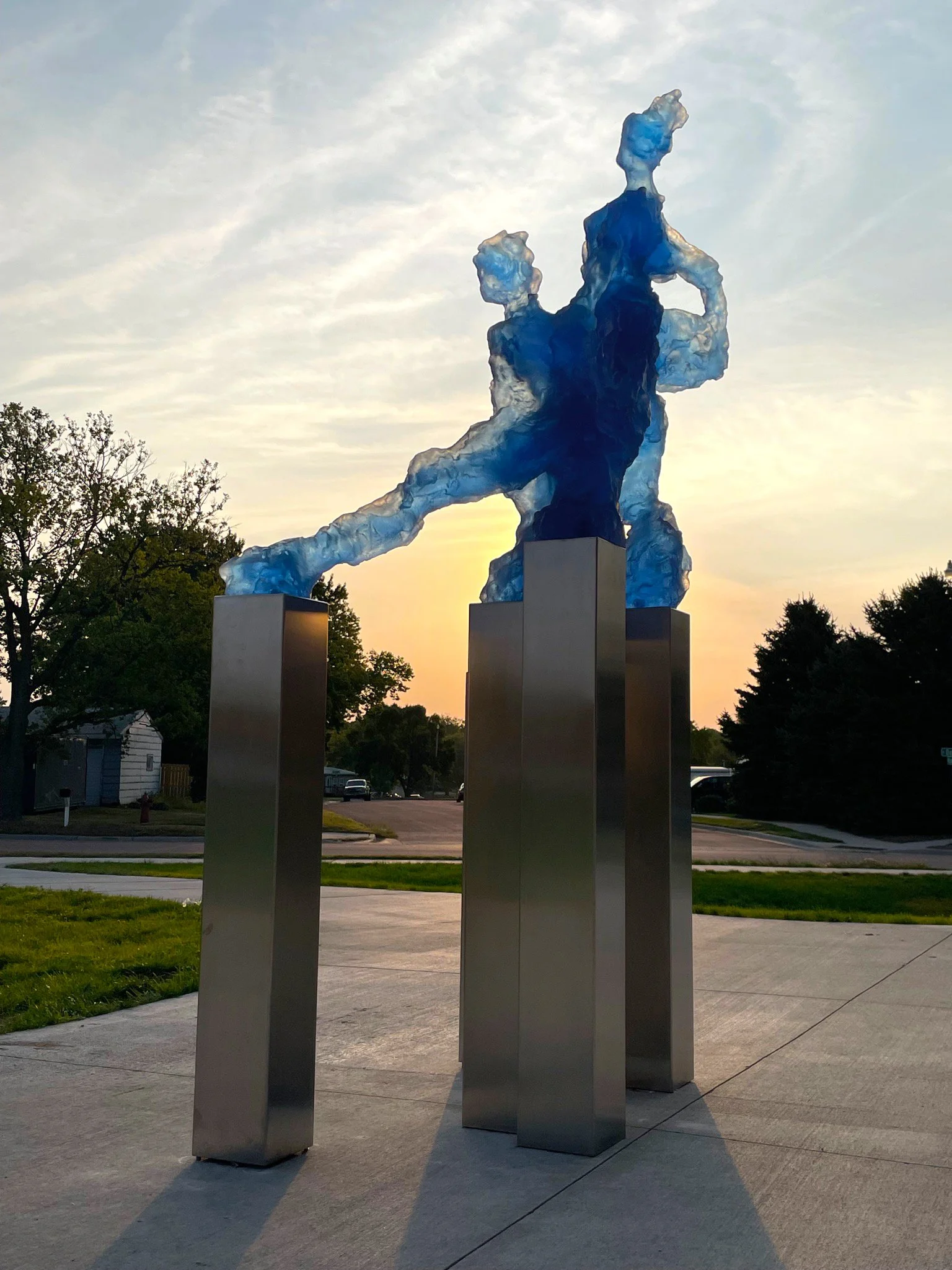 Sculpture of a group of translucent blue figures standing on reflective steel bases during sunset with trees and a street in the background.