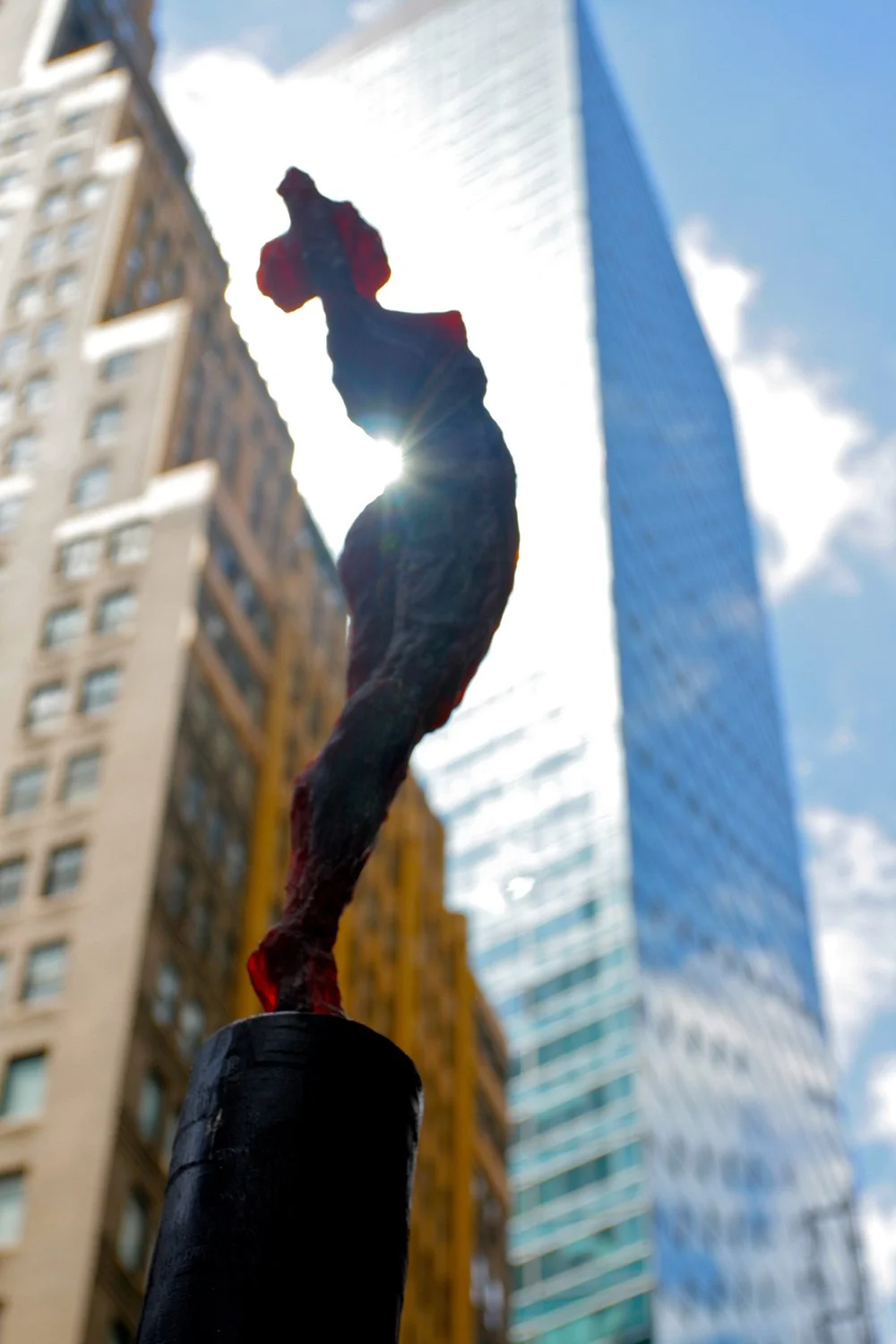 Close-up of a stylized sculpture of a stretching female in front of tall modern buildings, with the sun shining behind.