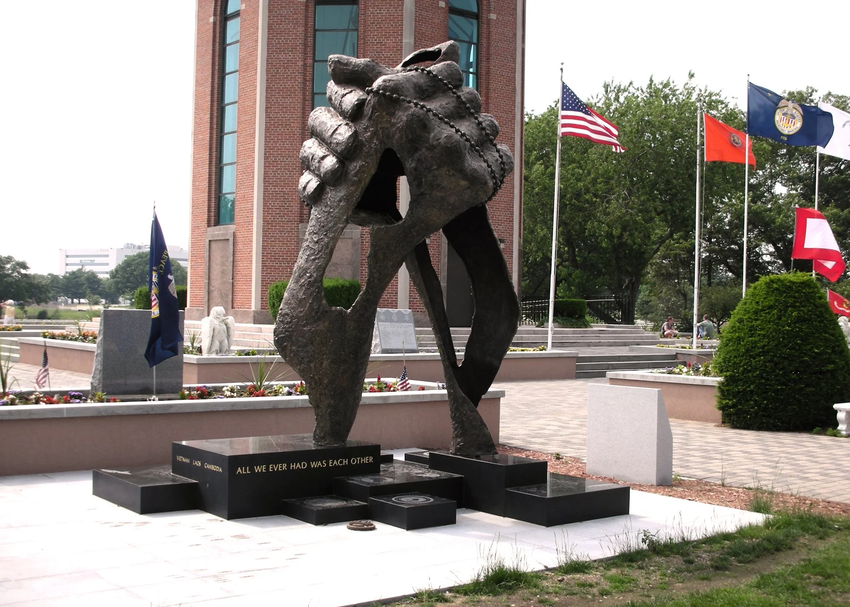 A bronze sculpture of two hands clasped together in symbol of Vietnam Veteran's brotherhood, set against a brick building and surrounded by flags, flowers, and small memorial plaques at a memorial site.