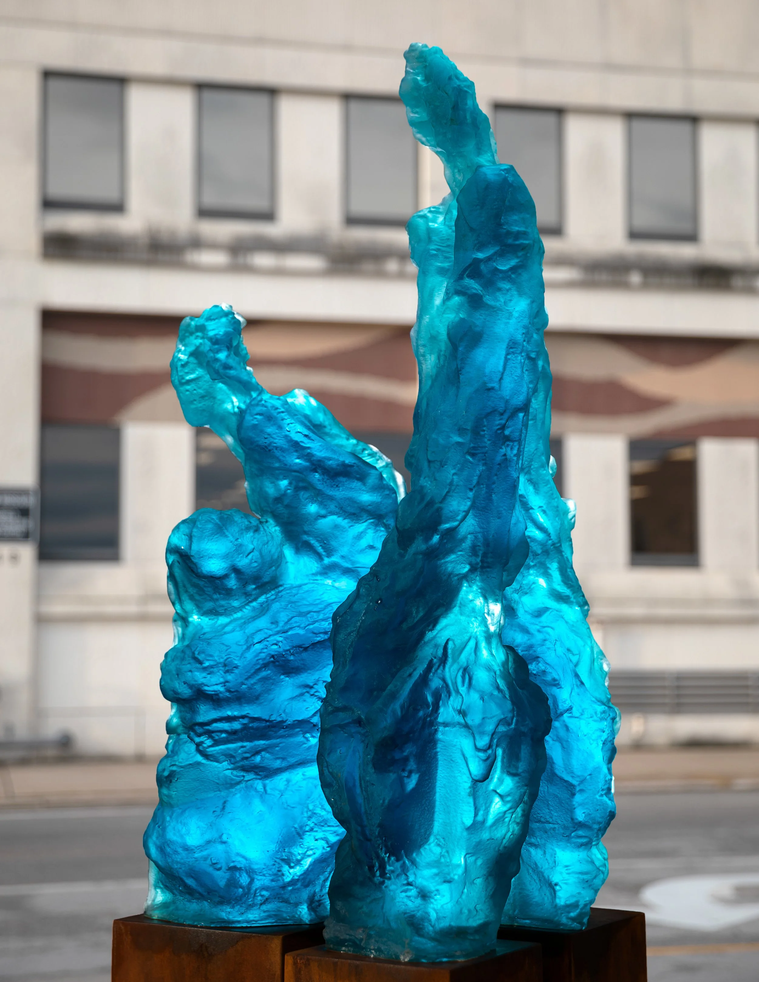 Blue glass sculpture resembling abstract flames, mounted on a wooden base, in front of a modern building with rectangular windows.