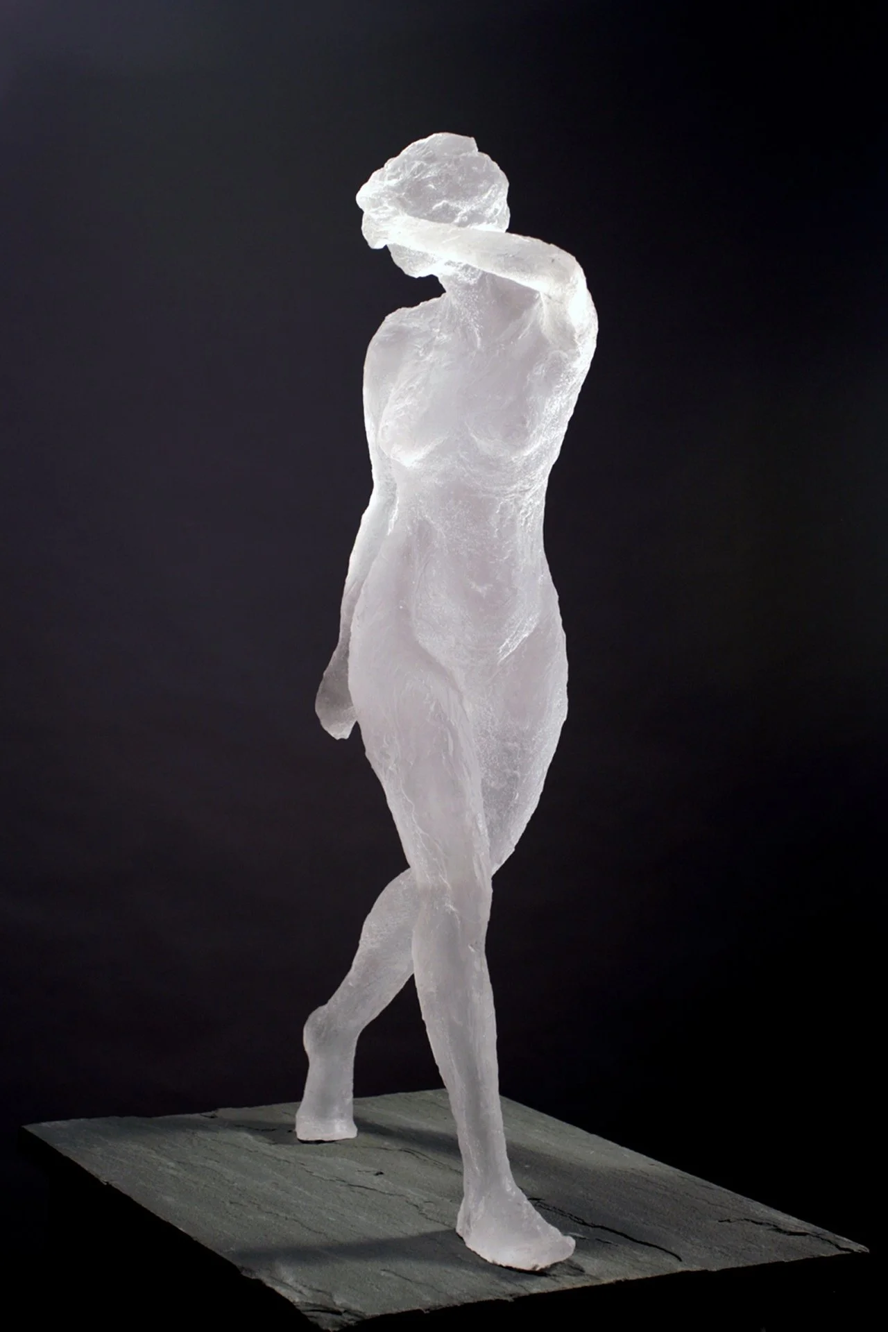 Ice clear sculpture of a woman standing on a base with one arm raised to her head against a black background.