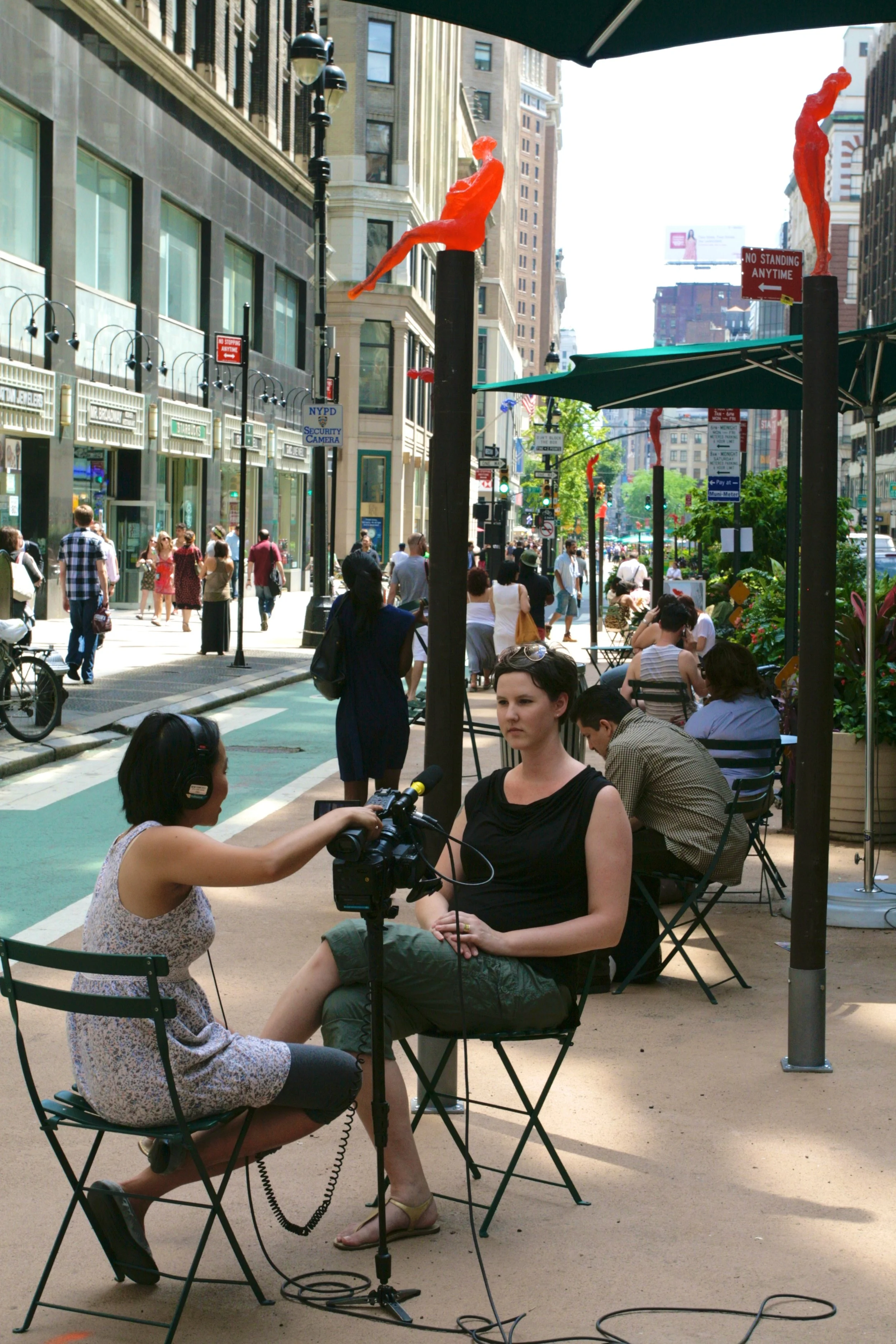 Two women are sitting and having a conversation on a city sidewalk during daytime, with one woman holding a microphone and camera for an interview in front of her. People are walking in the background, and there are tops of some buildings and street 