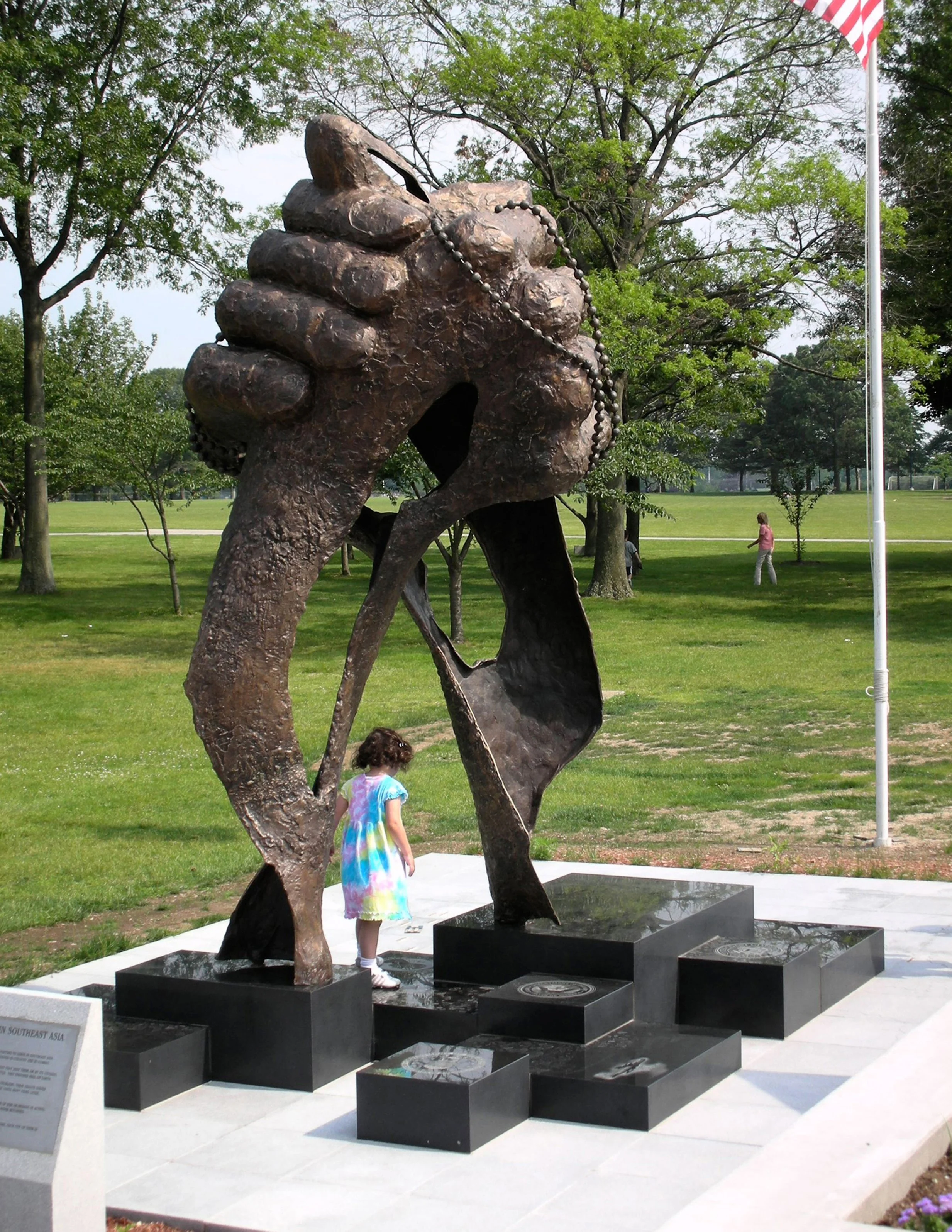 A large metal sculpture of a hand holding dog tags, situated outdoors on a black platform with surrounding trees and grass. Several children are nearby, with one girl in a colorful tie-dye dress touching the sculpture.