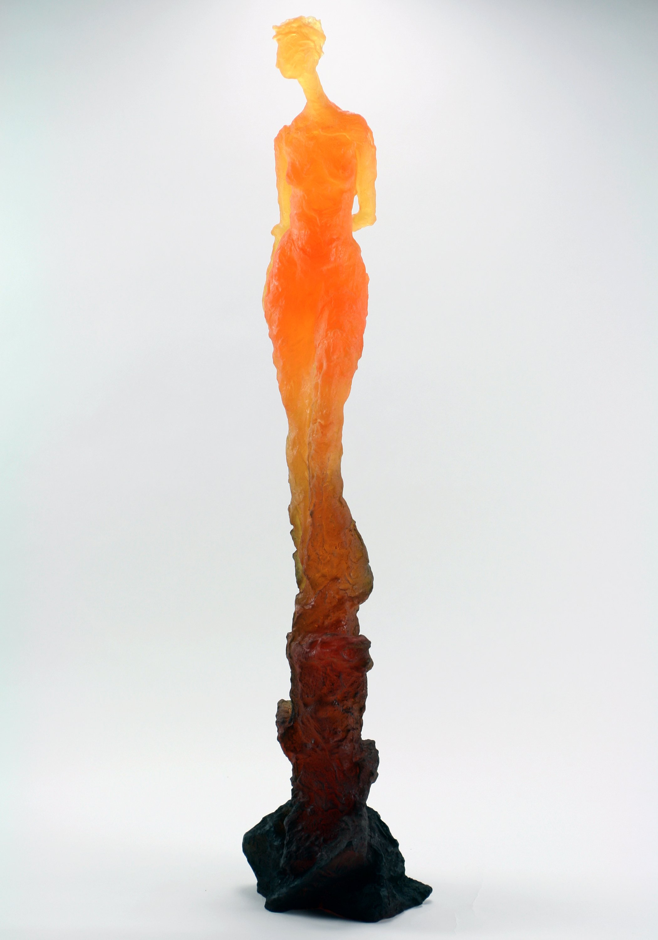 A tall, abstract sculpture resembling a human figure with an elongated body and head, in shades of orange, yellow, and red, standing on a dark, rocky base against a plain light background.
