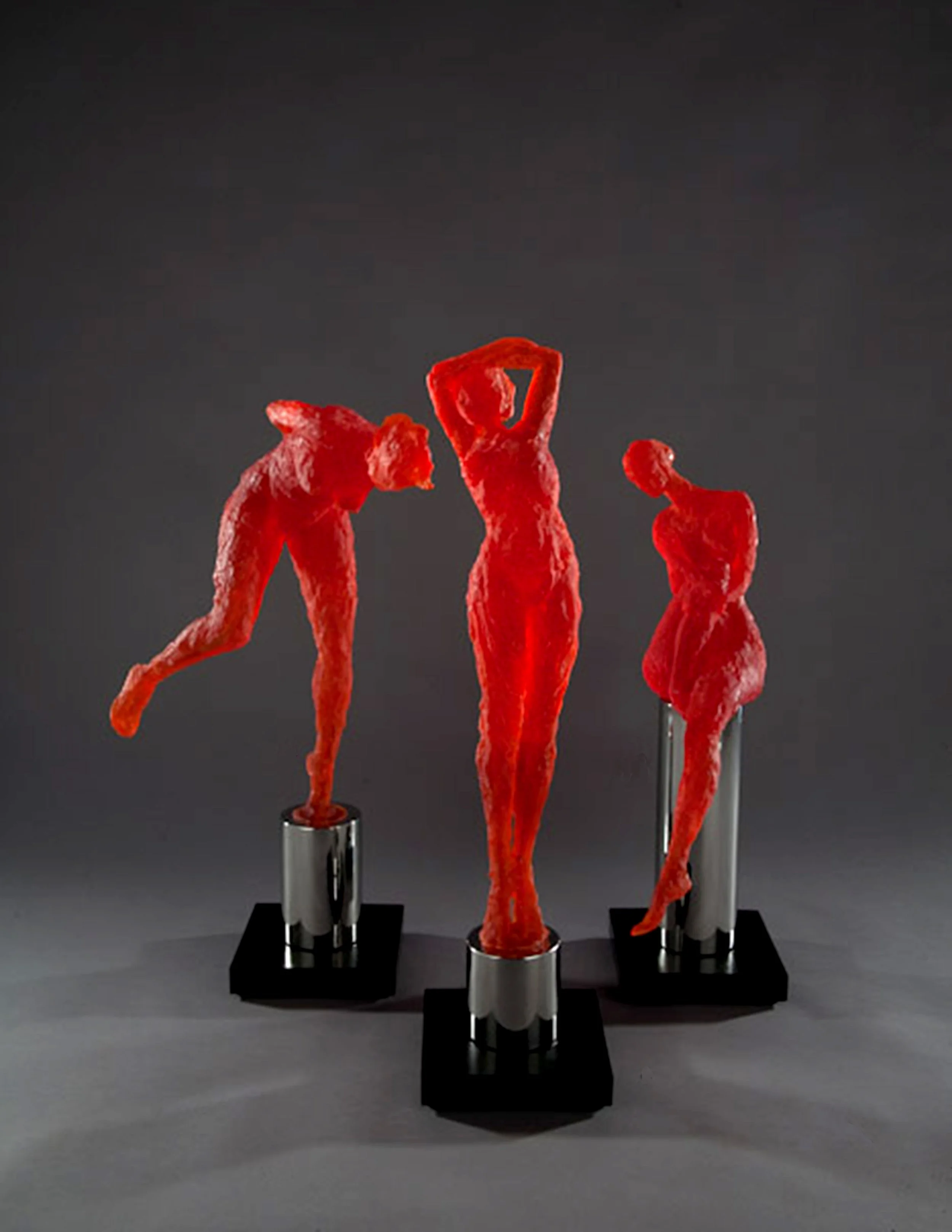 A display of four red, abstract sculptures resembling elongated human figures, mounted on black bases against a dark gray background.